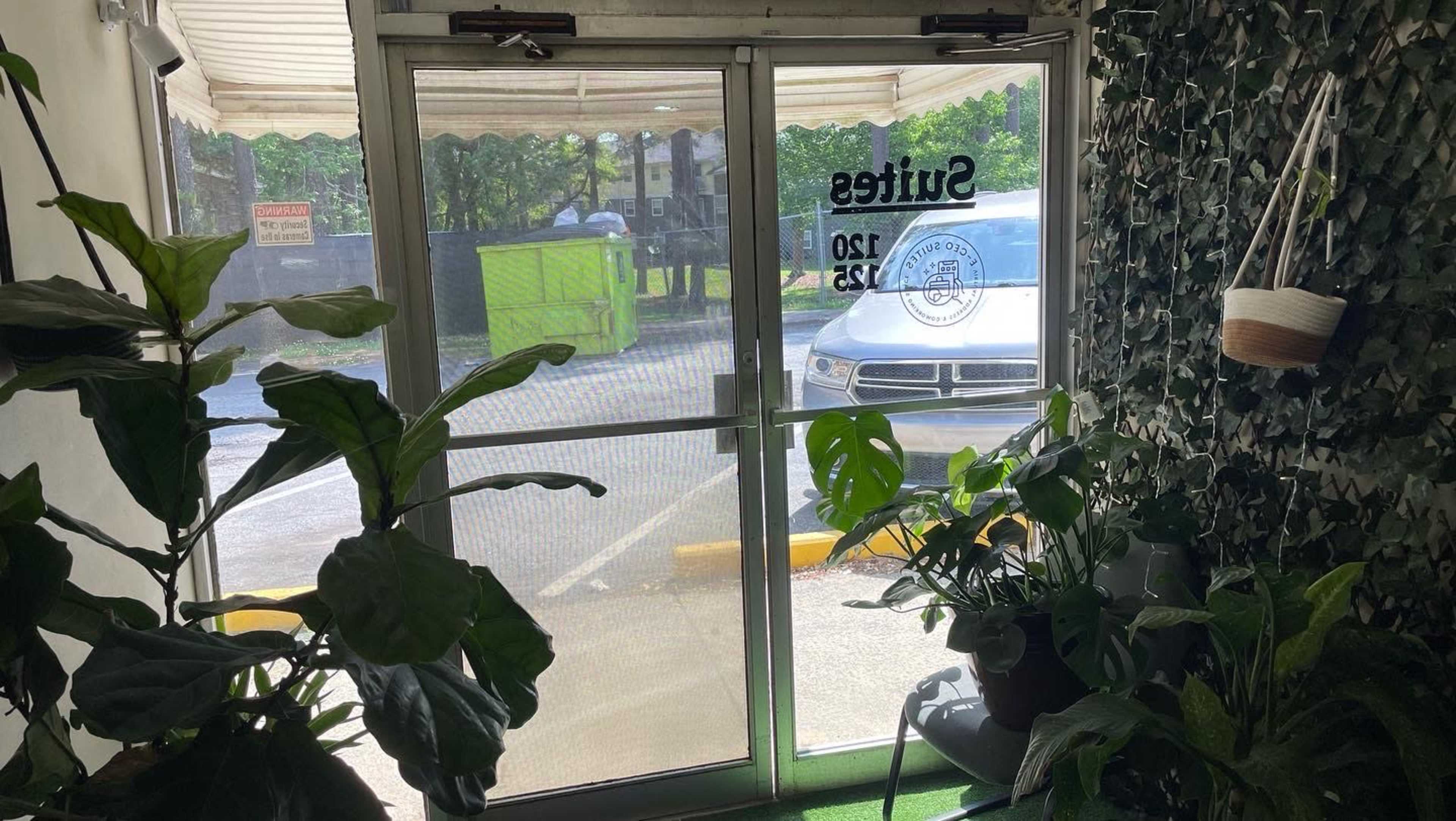 The image shows a glass entrance door with a leafy wall on one side and a view of a parking lot through the door.