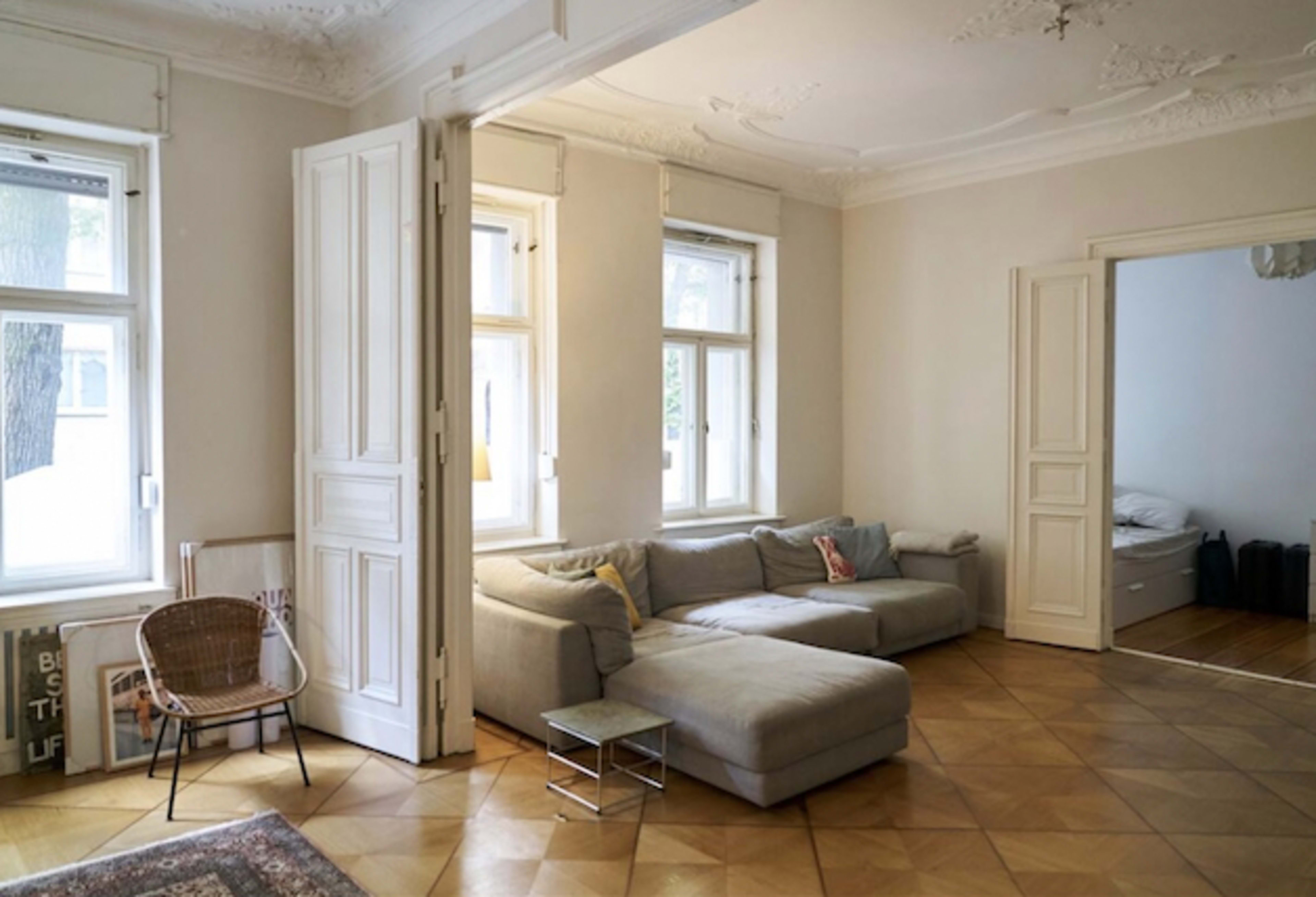 A spacious living room features a gray sectional sofa, a small metal side table, and a wicker chair beside large windows that allow natural light to fill the space.