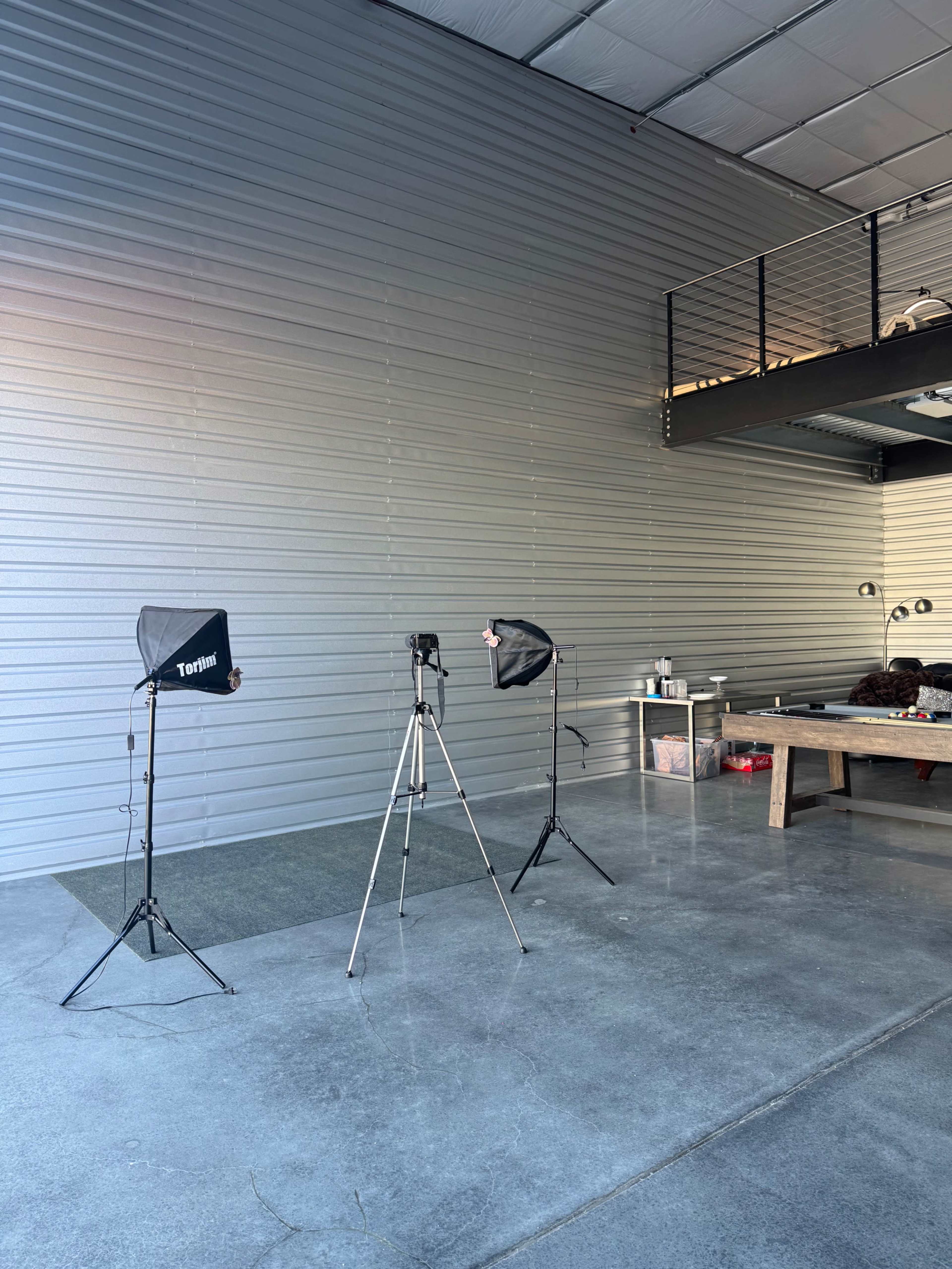 The image shows a spacious room with a gray wall, two photo lights on stands, and a camera mounted on a tripod, along with a table in the background.