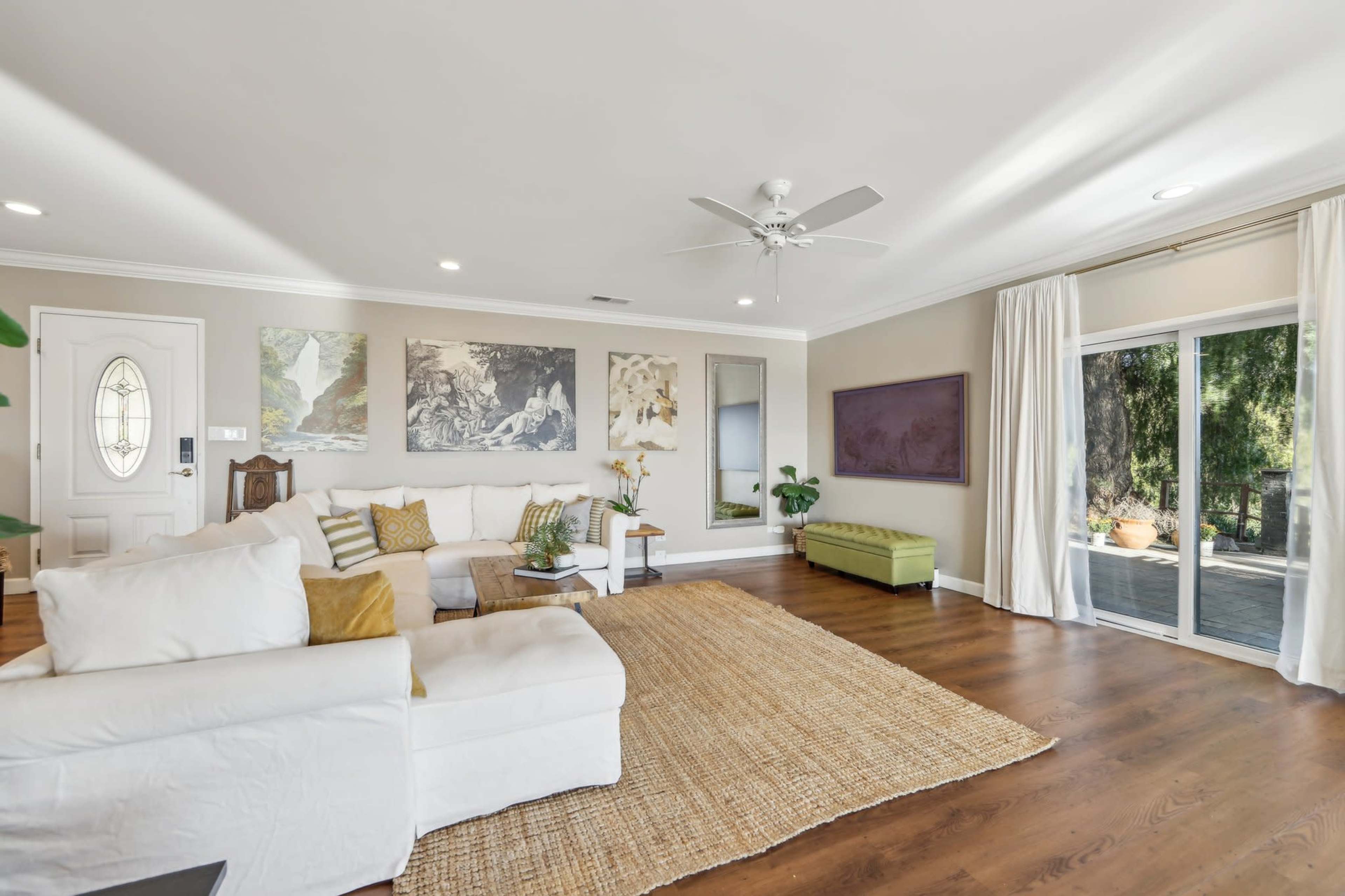The image shows a bright living room with a white sectional sofa, a large area rug, artwork on the walls, and glass doors leading to an outdoor patio.