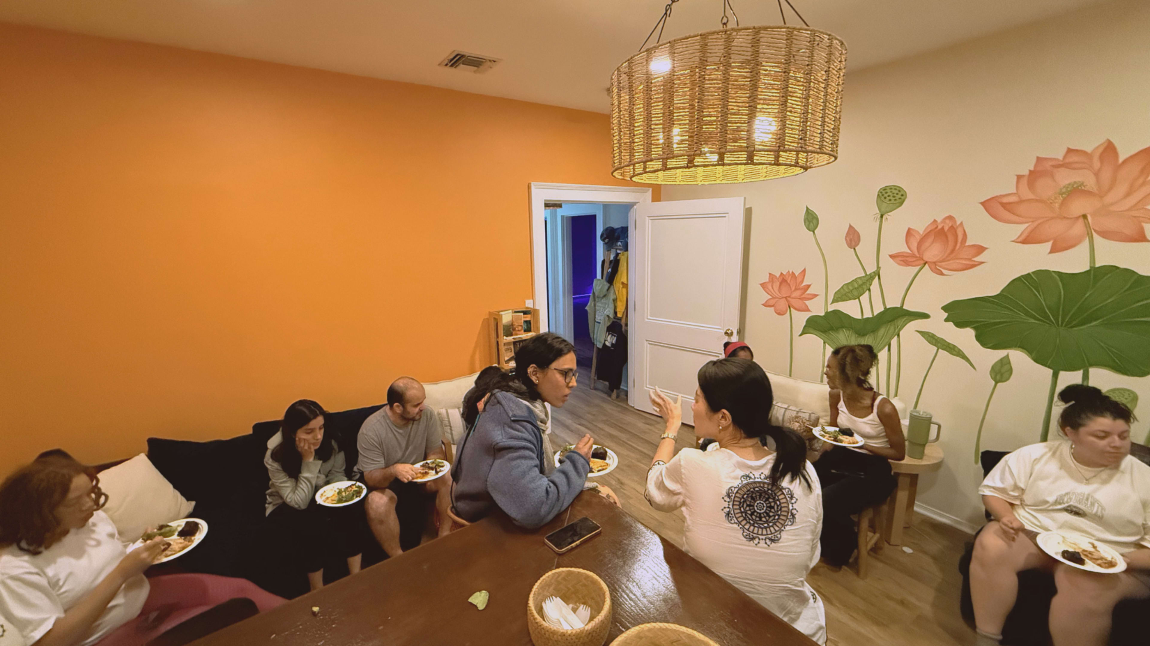 A group of people are seated around a dining table and couch, enjoying food and engaging in conversation in a brightly colored room with a floral mural.