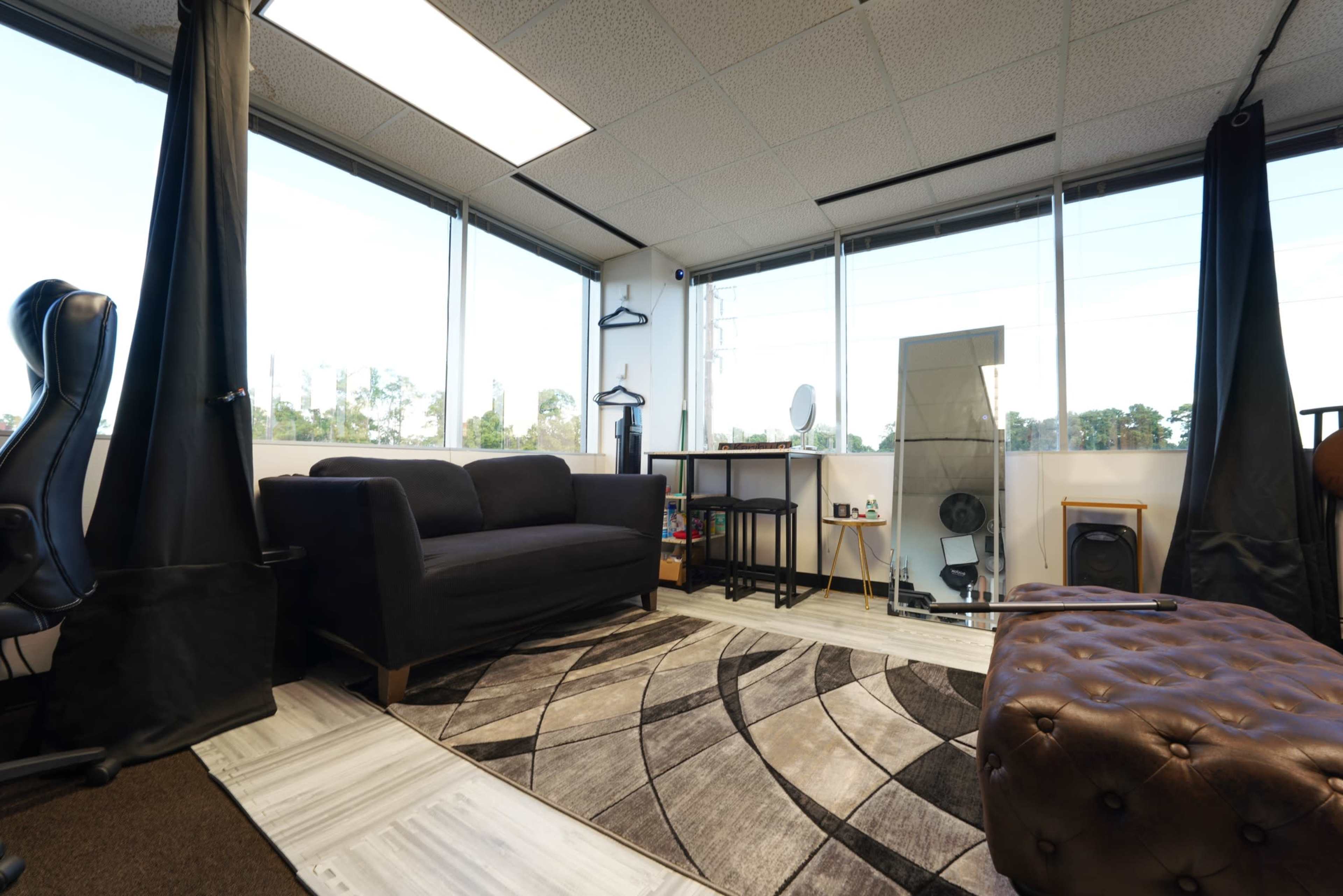A spacious office or studio with large windows, a black sofa, a patterned rug, a wooden shelf, and a leather tufted ottoman.