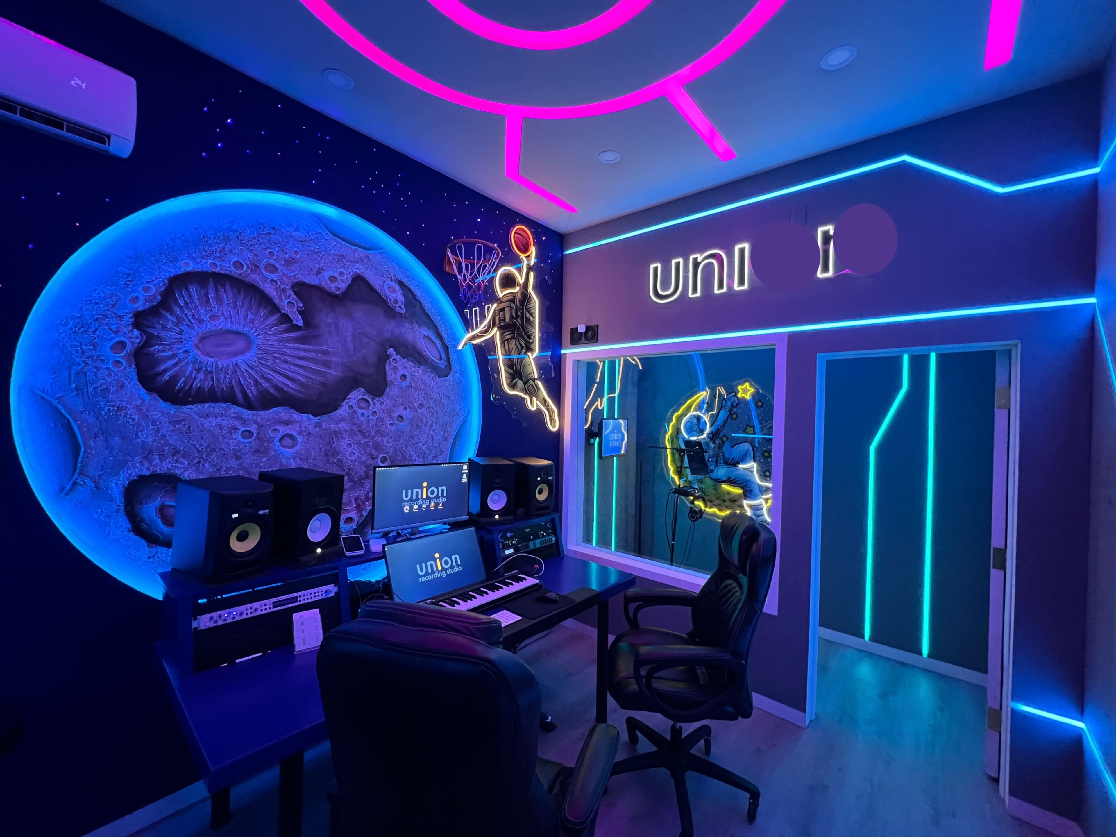 The image shows a modern music studio featuring a dark wall mural of a planet, neon lighting, and an illuminated sign that reads "uni."