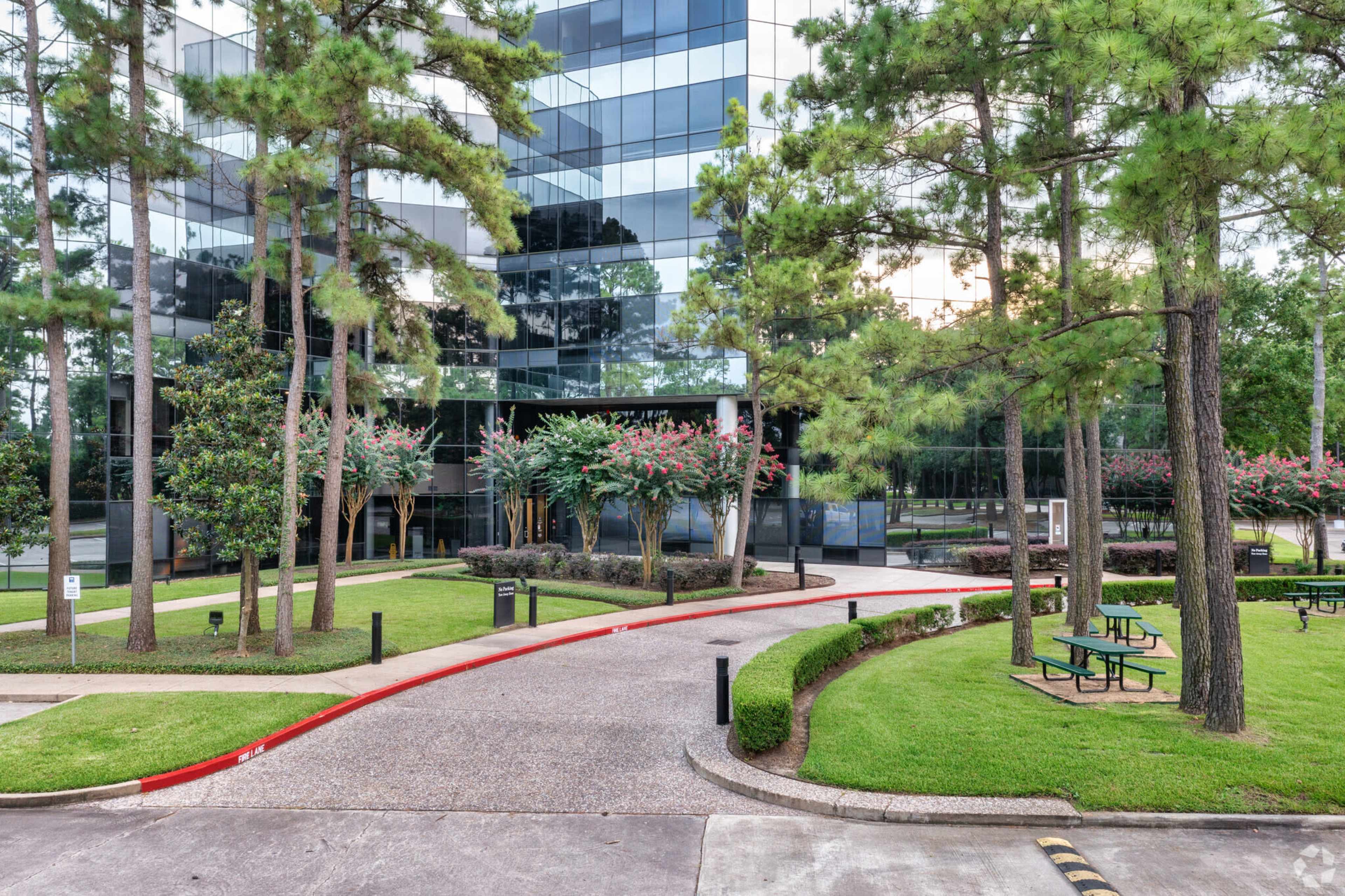 The image depicts a landscaped area with a winding path surrounded by trees and shrubs, leading to a modern glass office building.