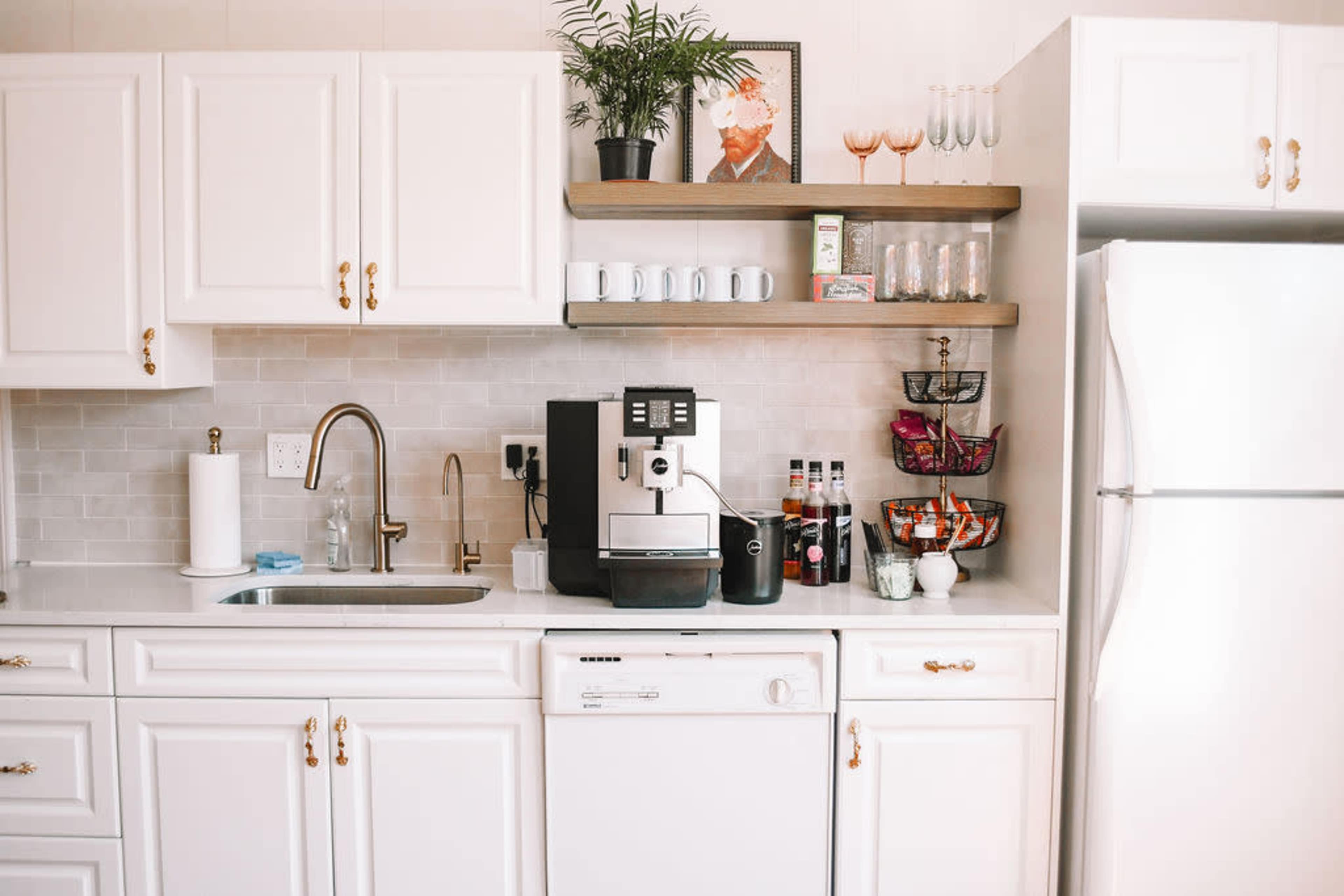 The image shows a modern kitchen with white cabinetry, a stainless steel refrigerator, an espresso machine on the counter, and various glassware displayed on open shelves.