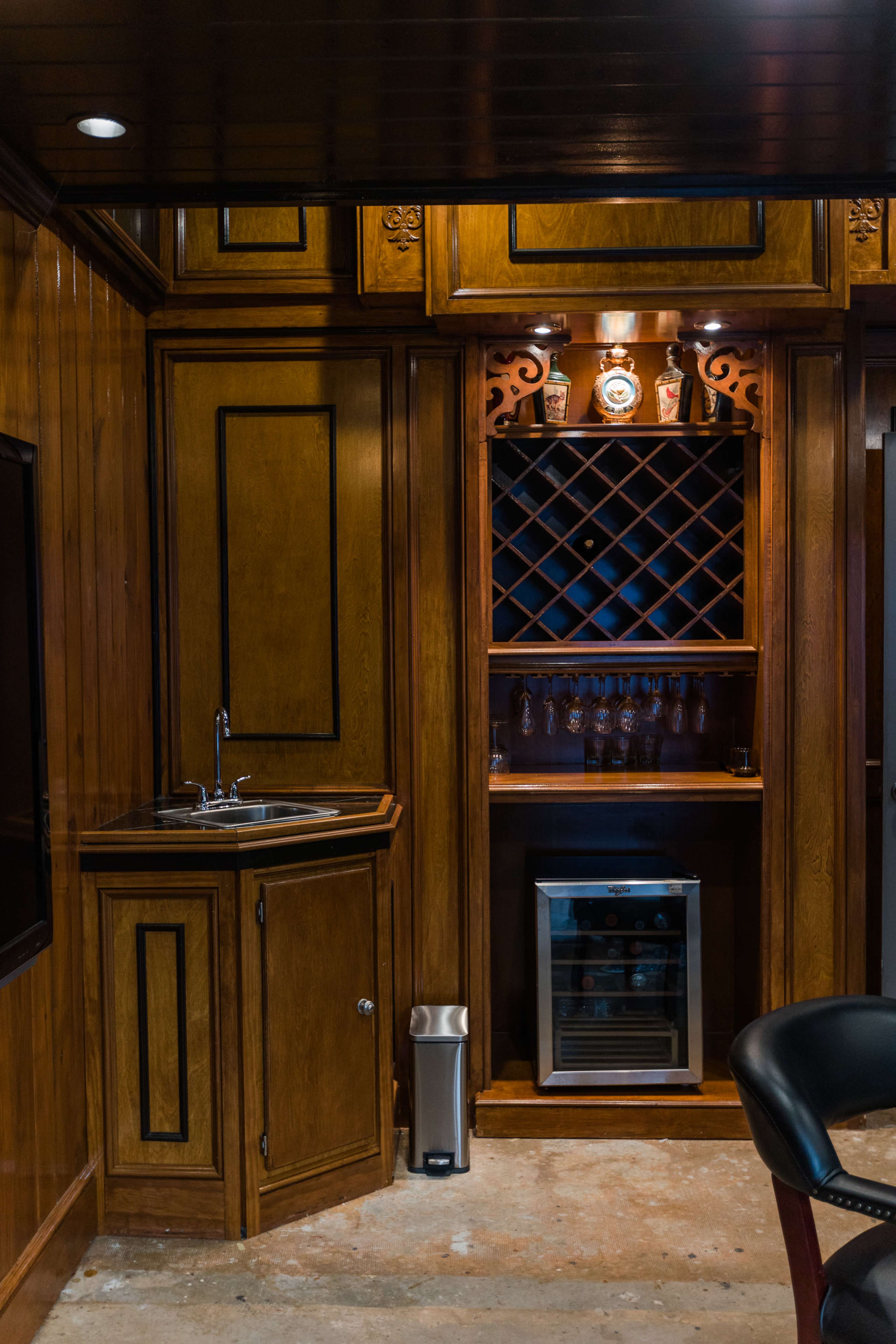 A wooden bar area with a sink, wine rack, and a small refrigerator in a room with wood-paneled walls.