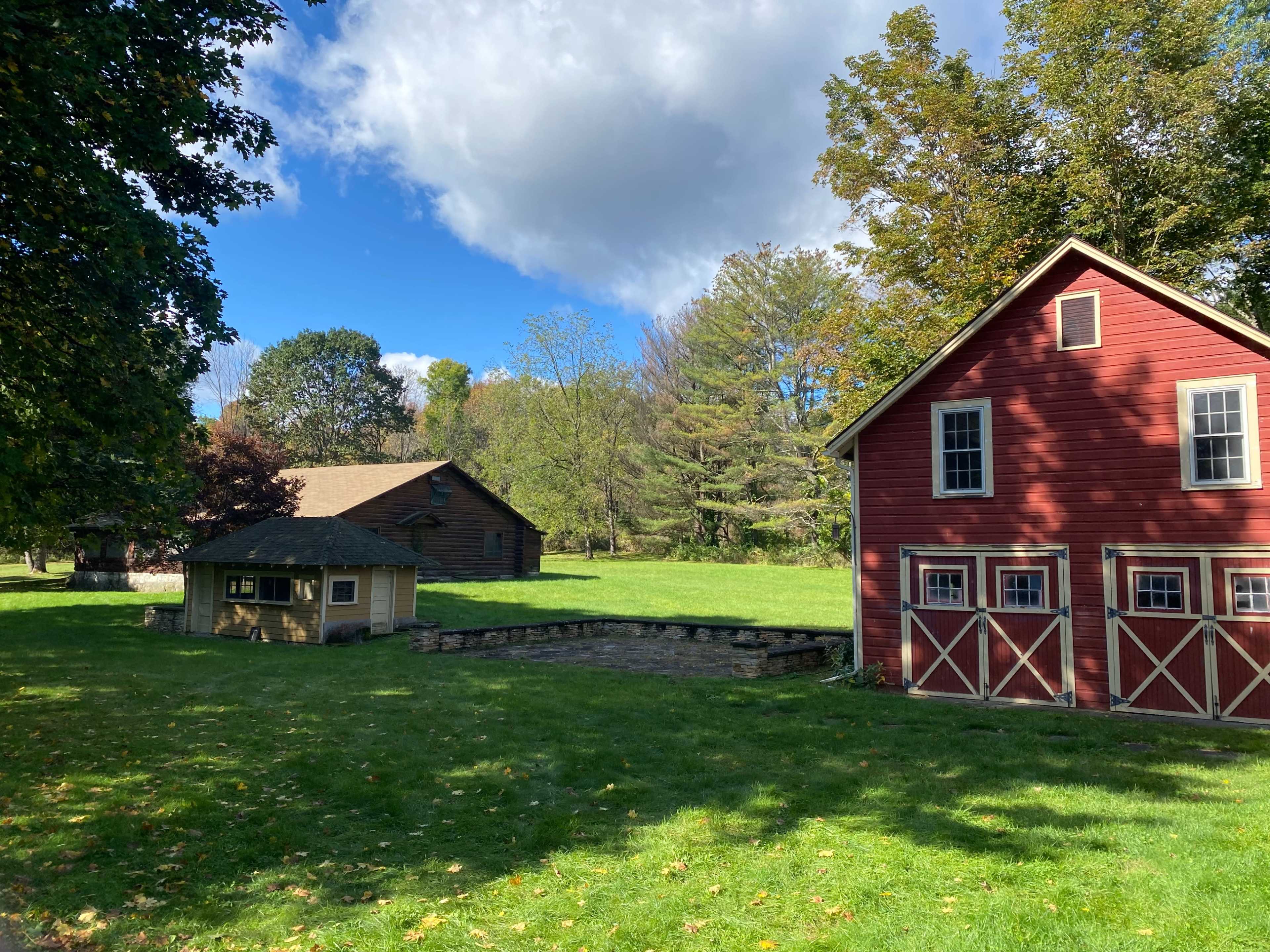 A red house with white trim next to a smaller building and a larger barn, all situated in a grassy area surrounded by trees under a partly cloudy sky.