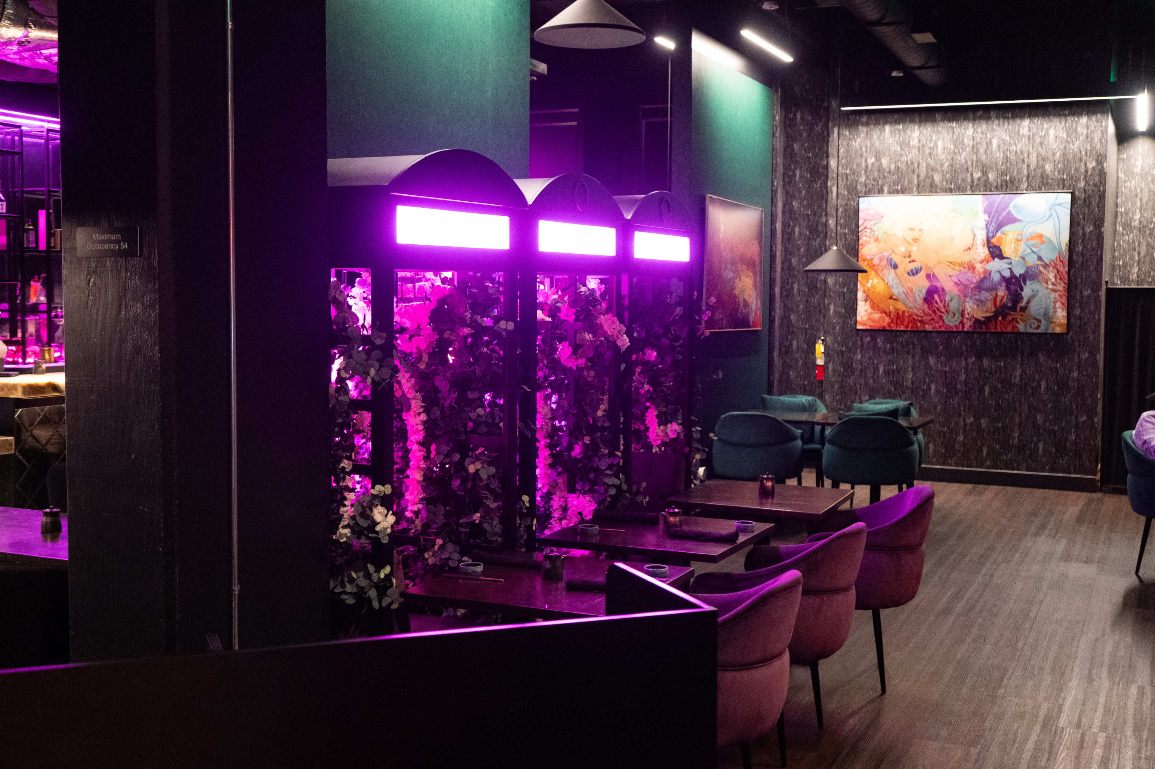 The image shows a modern restaurant interior featuring purple lighting, floral decor, and stylish seating arrangements.
