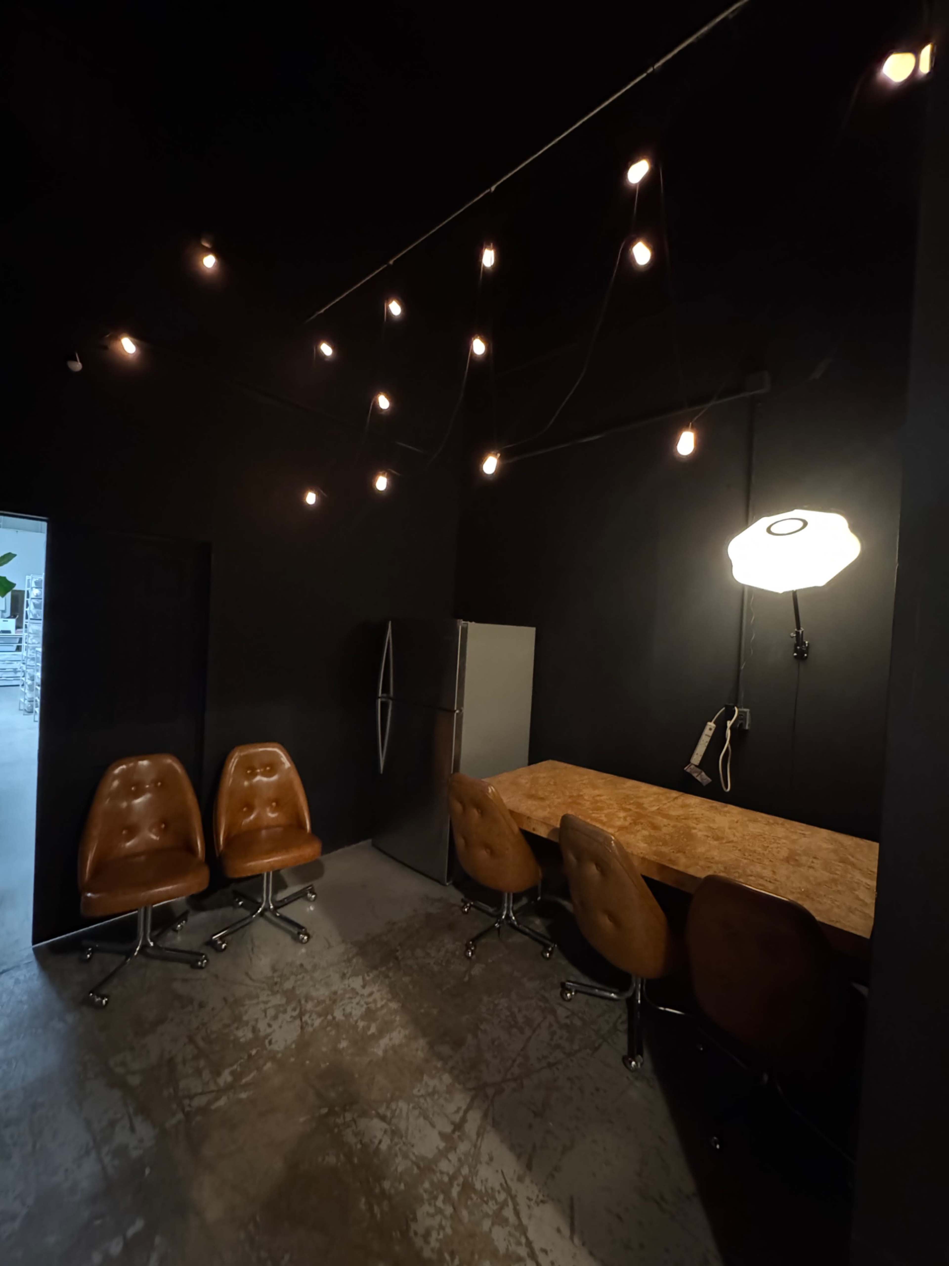 The image shows a dimly lit room with a wooden table and four brown swivel chairs, along with a refrigerator and hanging light bulbs above.