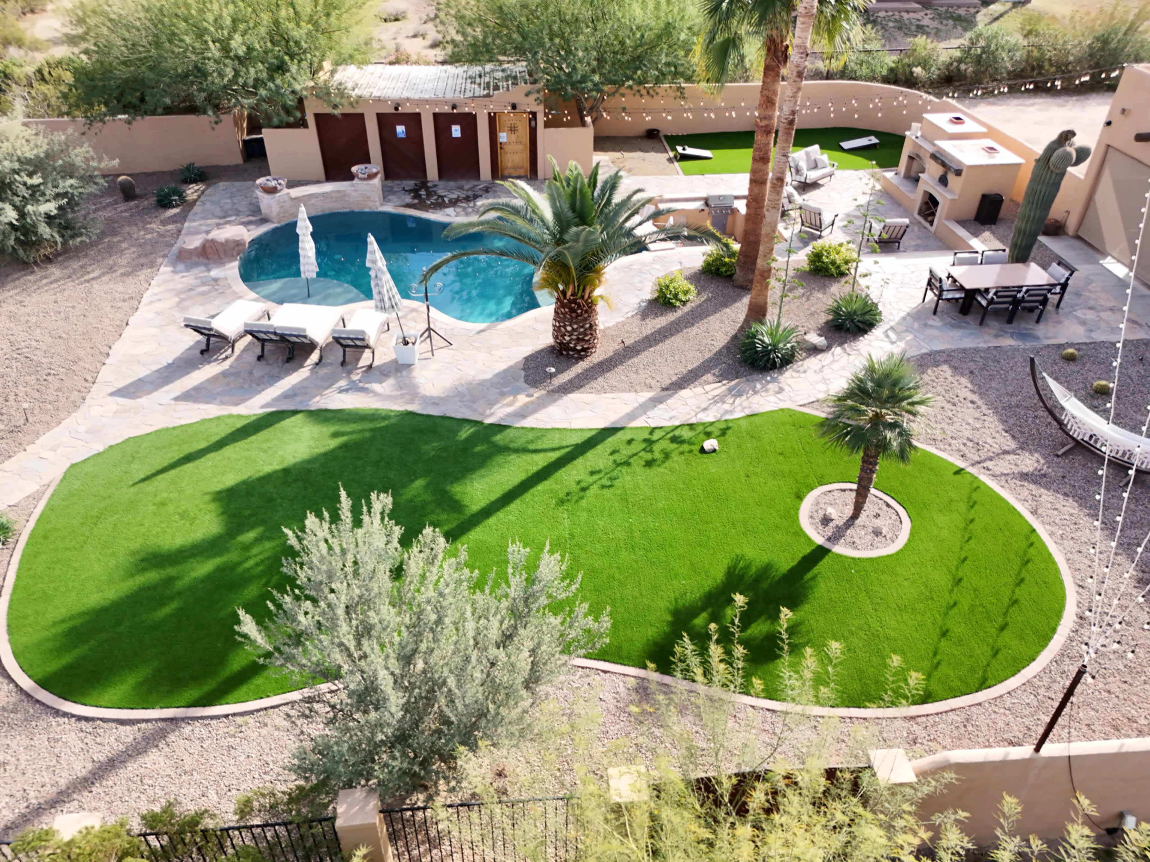 The image shows a landscaped backyard featuring a swimming pool, lounge chairs, palm trees, and a barbecue area surrounded by artificial grass.