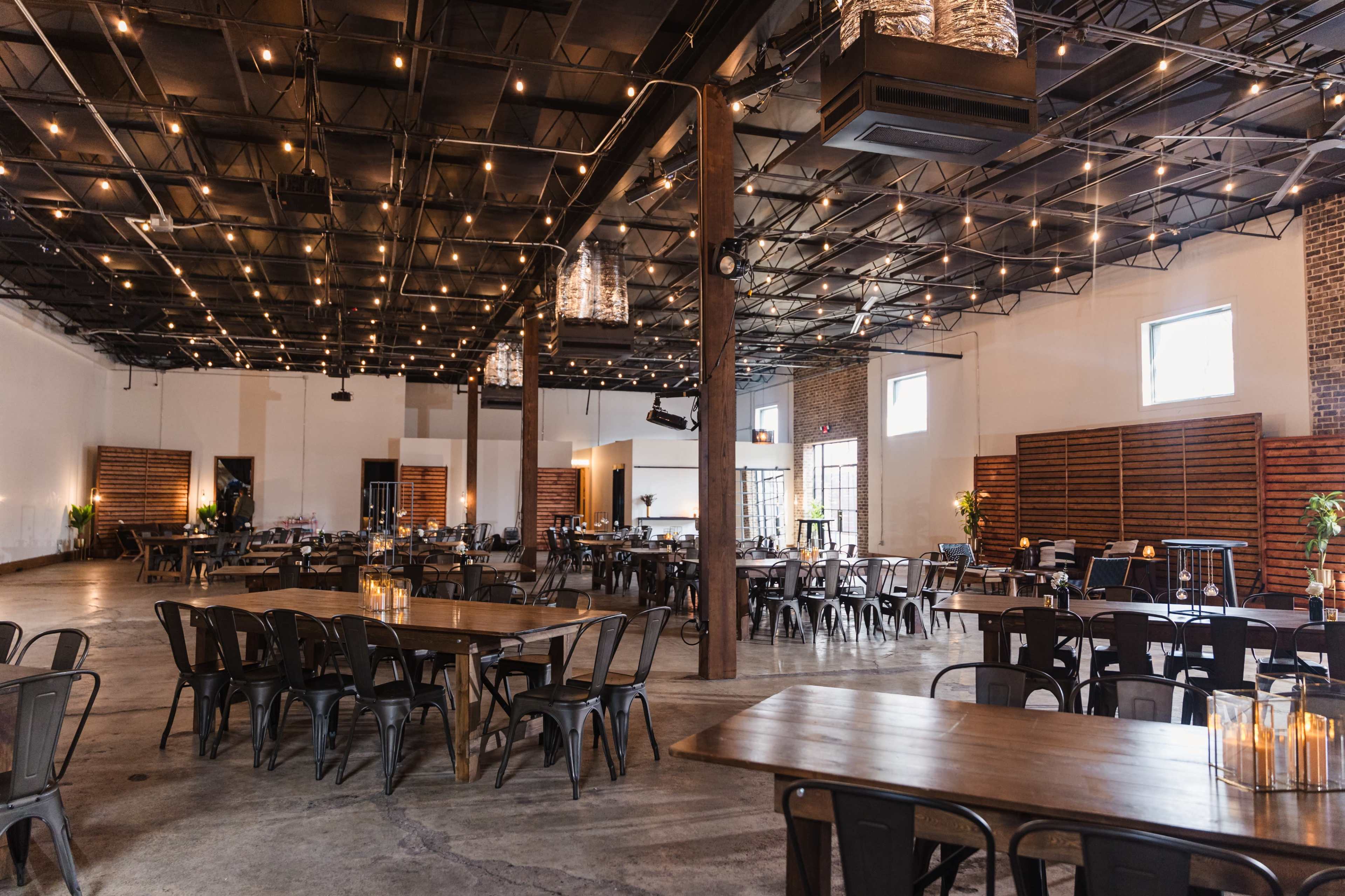 The image shows a spacious event venue with rows of tables and chairs, illuminated by string lights hanging from the ceiling.