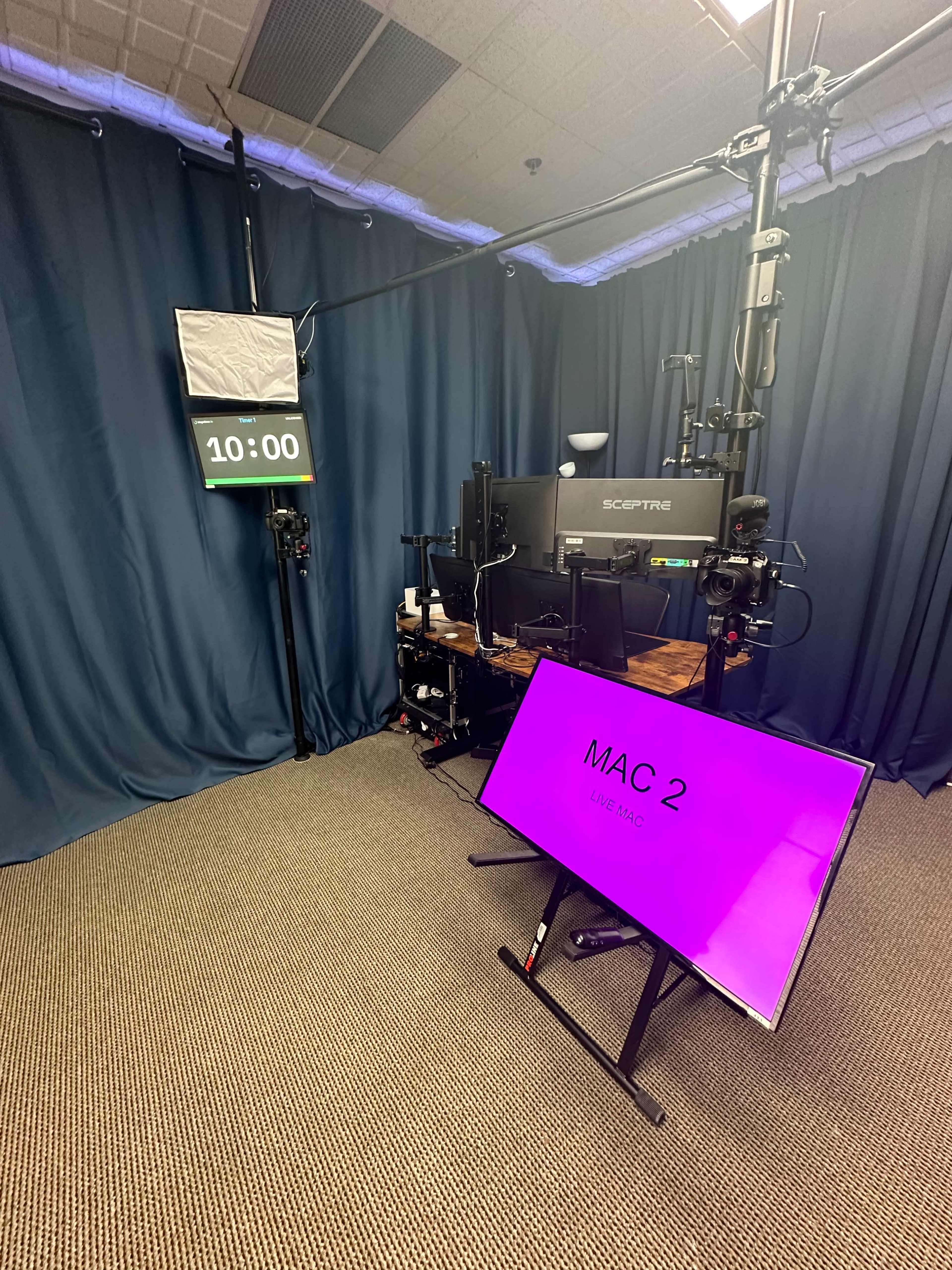 A recording setup featuring a large monitor displaying "MAC 2," a digital clock set to 10:00, and black curtains in the background.