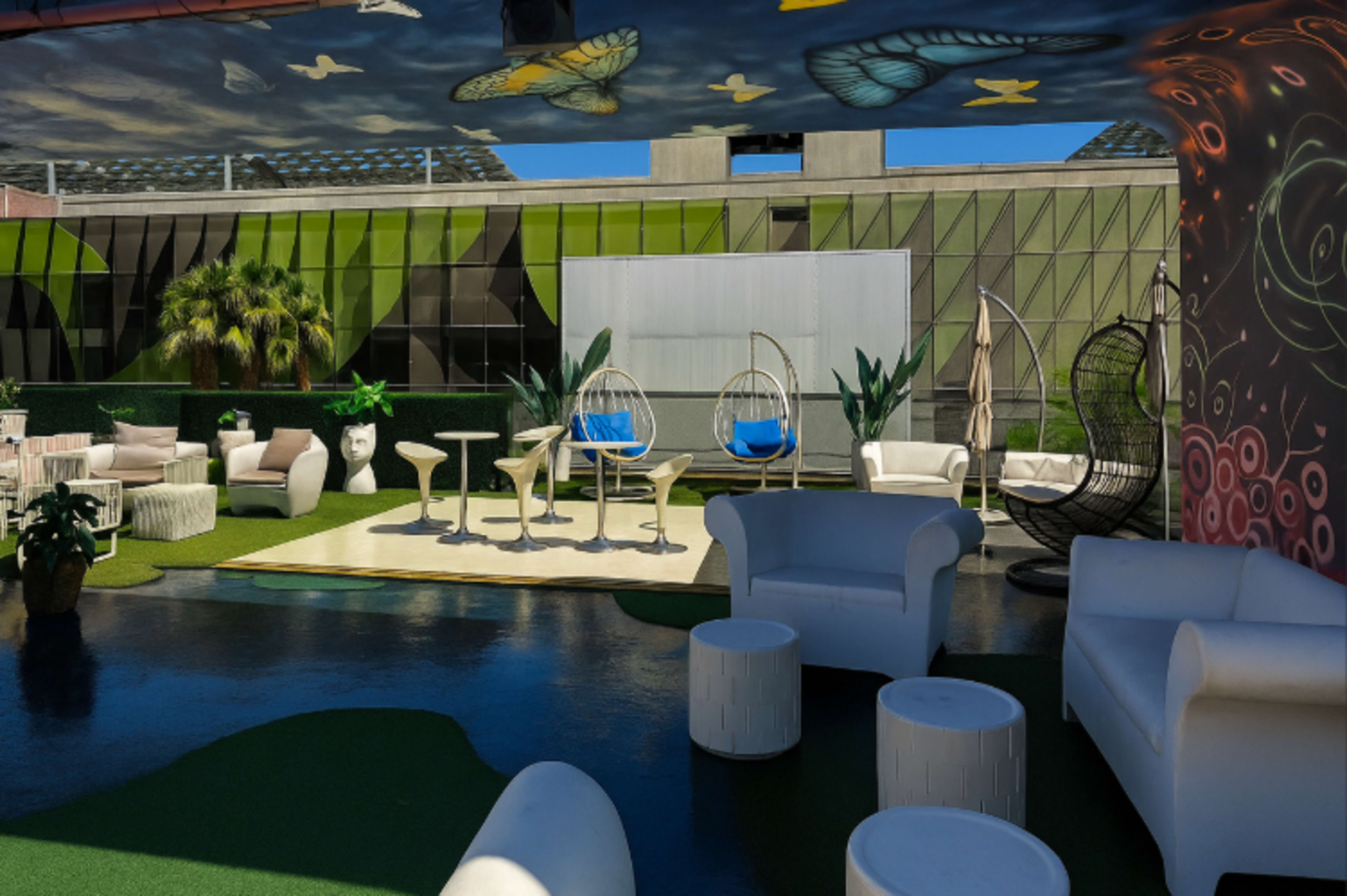 The image shows a modern outdoor lounge area featuring hanging chairs, white furniture, and green landscaping under a decorated ceiling.