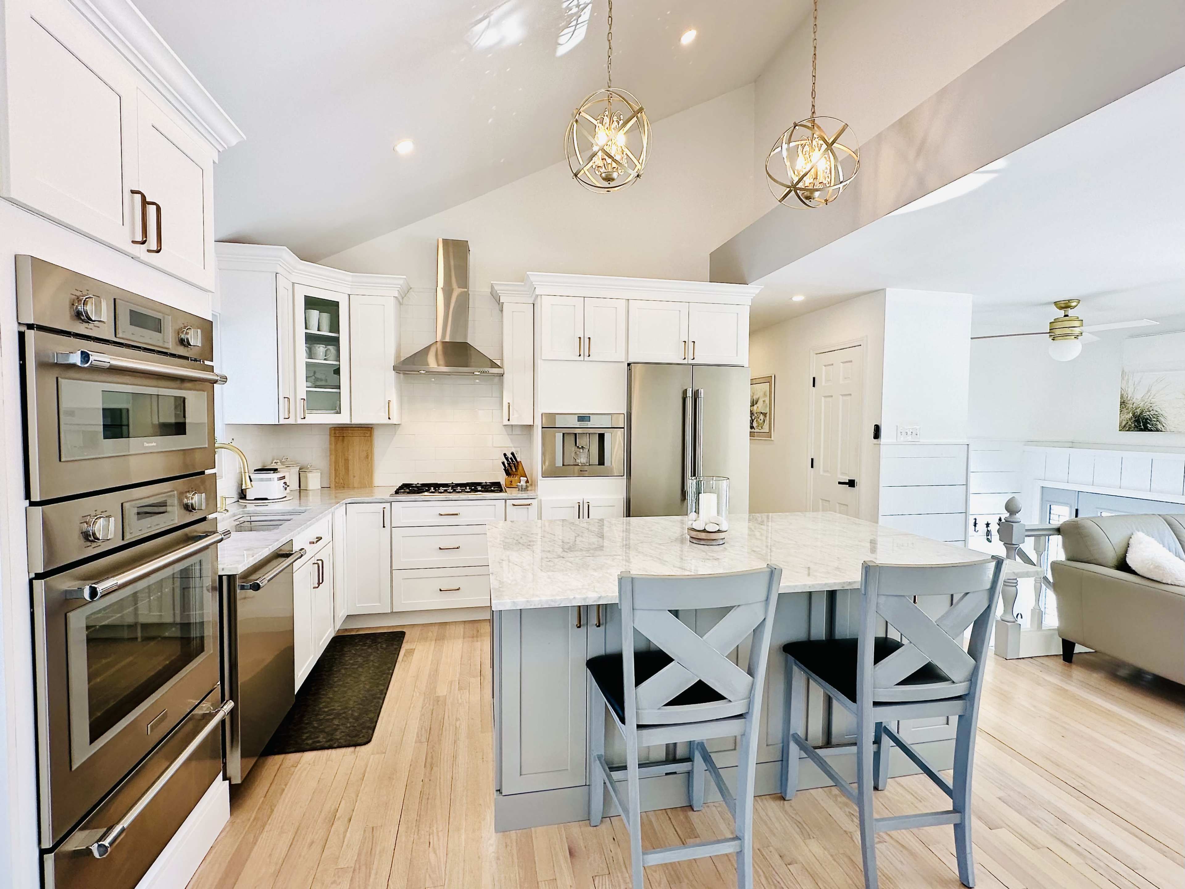 The image shows a modern kitchen with white cabinetry, stainless steel appliances, a marble countertop, and two gray bar stools at the island.