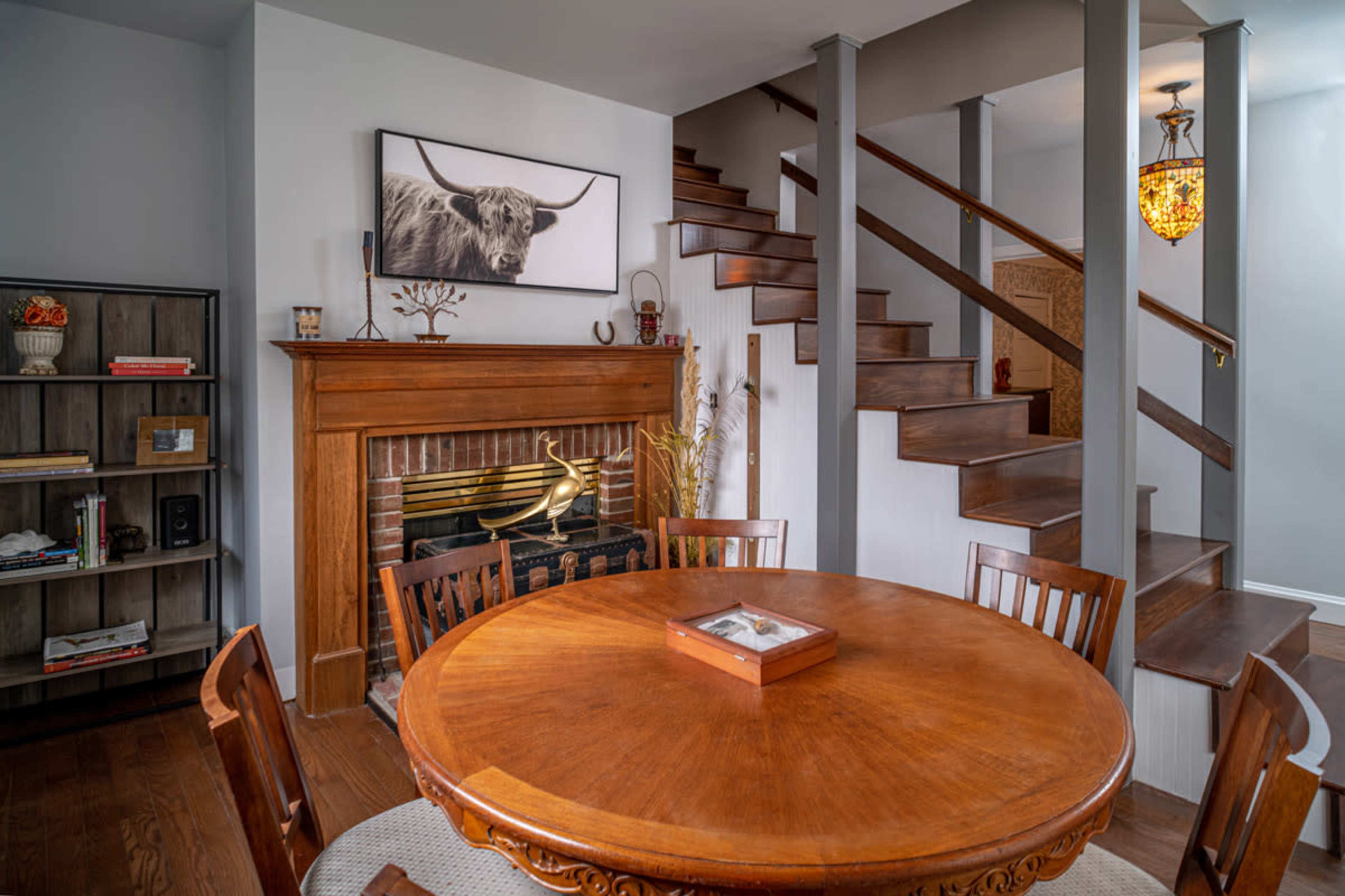 The image shows a dining area featuring a round wooden table with chairs, a decorative fireplace, and a staircase leading to an upper level.
