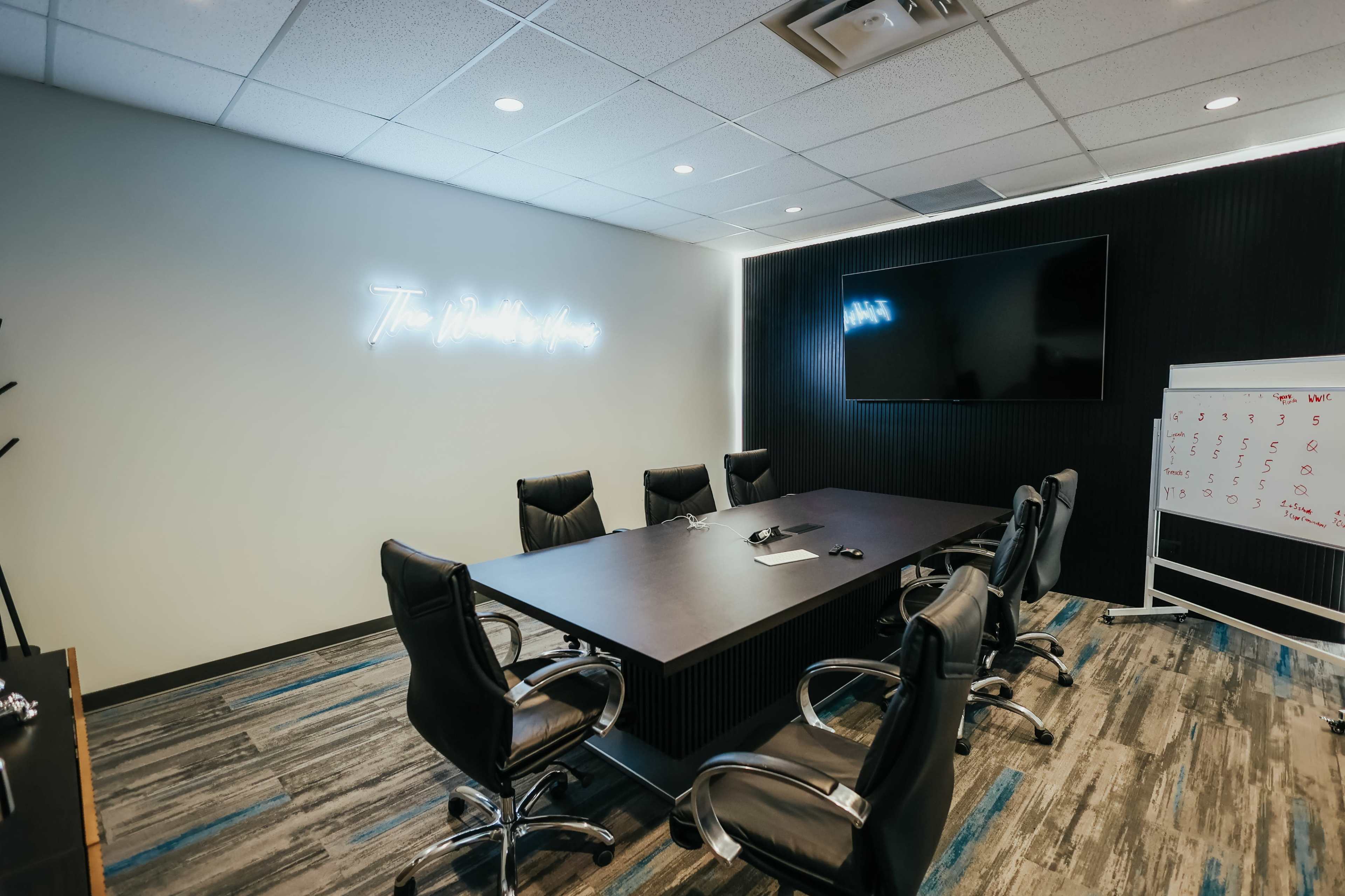A modern conference room features a large table with eight chairs, a television mounted on the wall, and a whiteboard beside it, all against a backdrop of sleek wall decor and carpeting.