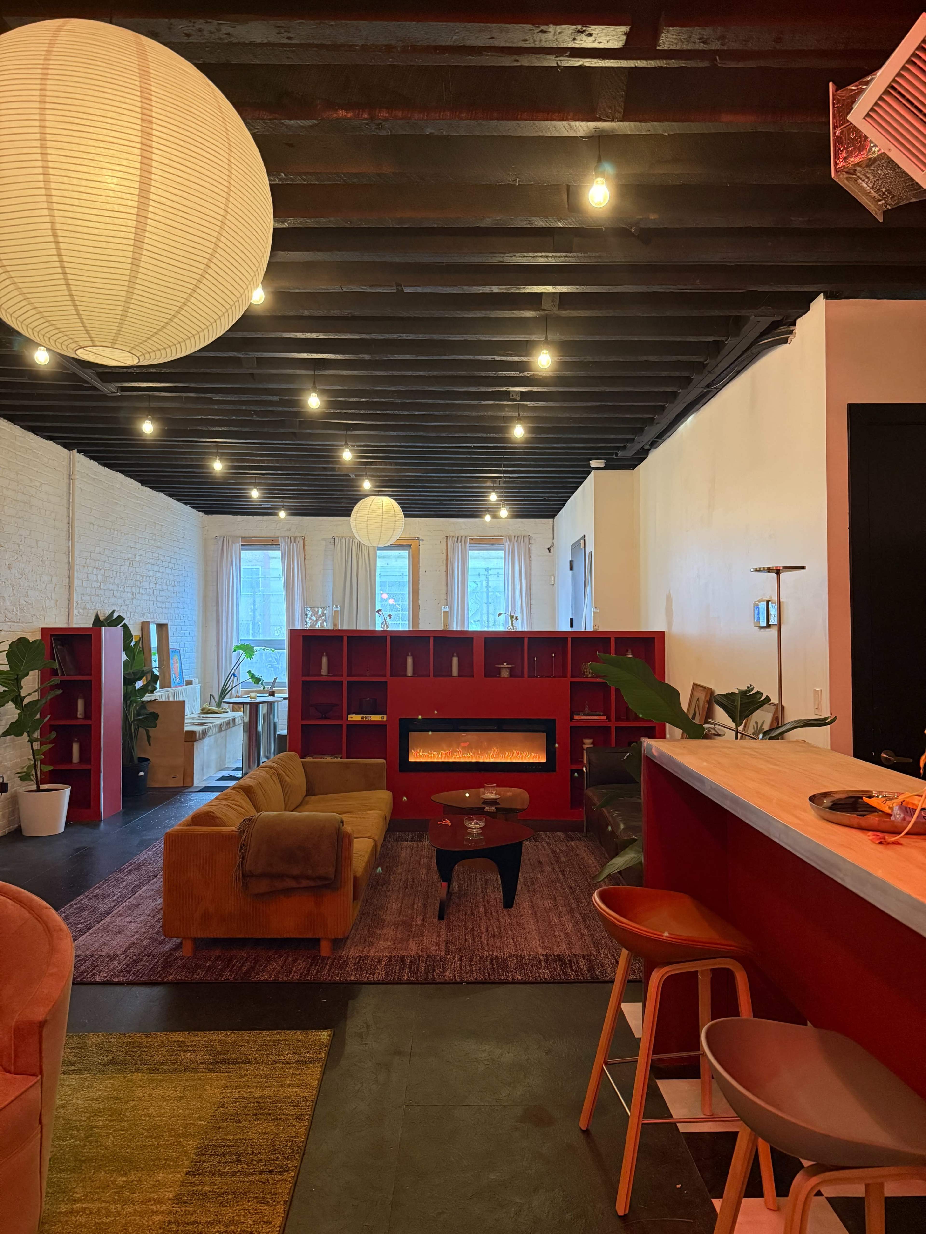 The image shows a modern, open living space featuring a cozy seating area, a red bookshelf, and a faux fireplace, with warm lighting and plants throughout.