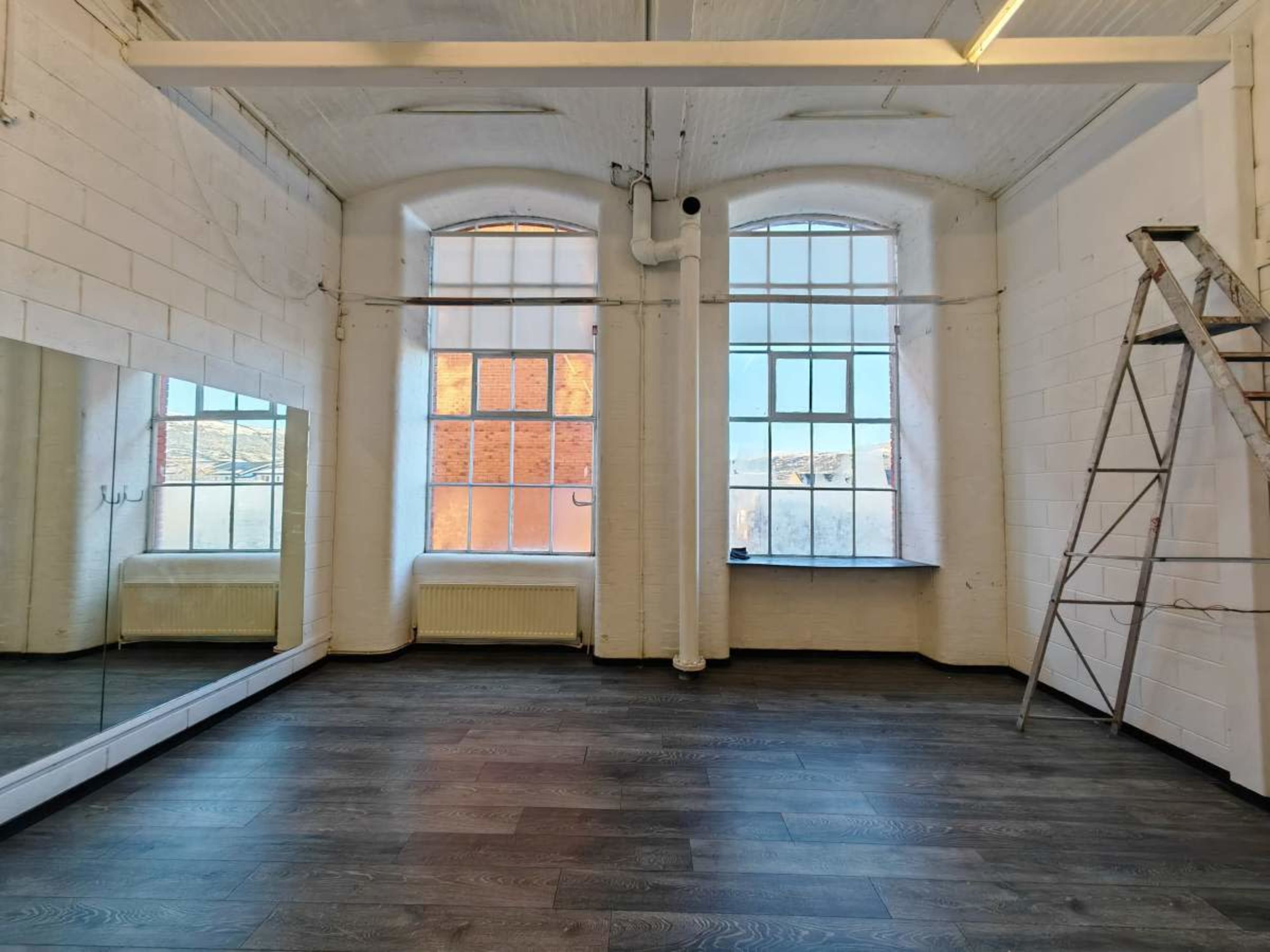 The image shows a spacious, well-lit room with large windows, a mirror on one wall, and a ladder leaning against another.