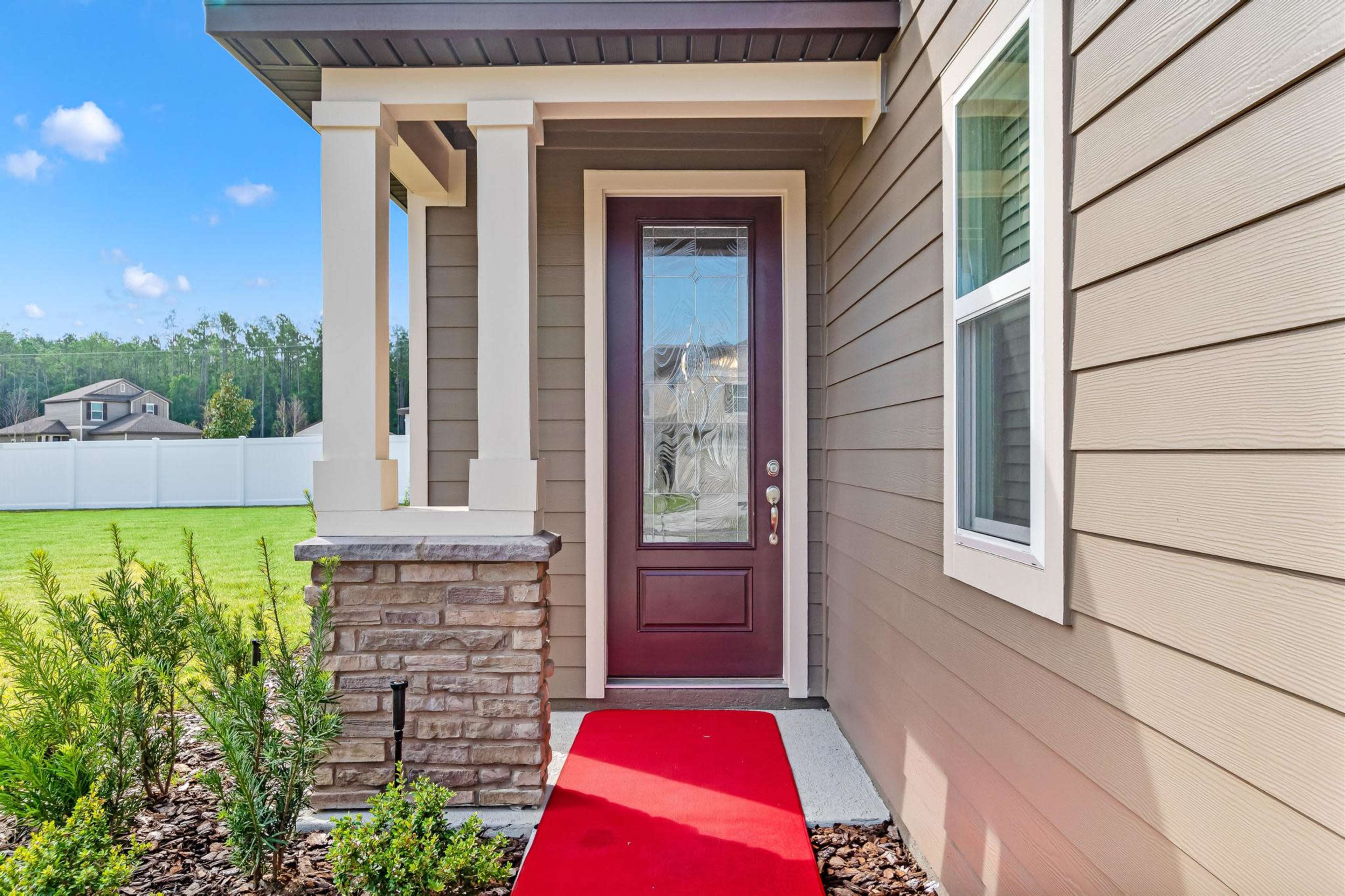 A front porch featuring a maroon door with a glass panel, flanked by light-colored siding and stone accents, and a red carpet leading to the entrance.