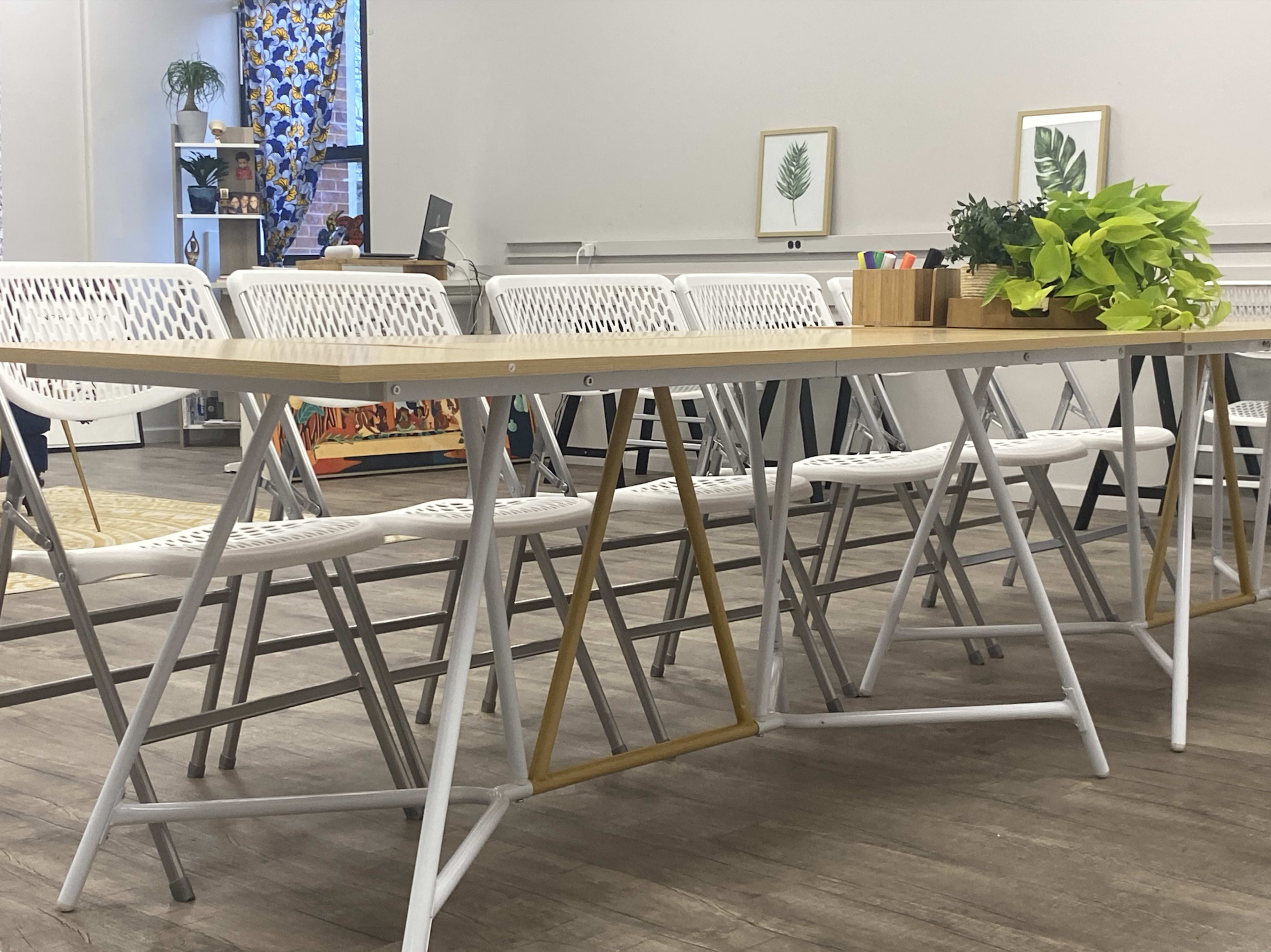 A row of white folding chairs arranged around a long wooden table in a bright workspace, with potted plants and framed artwork on the walls.