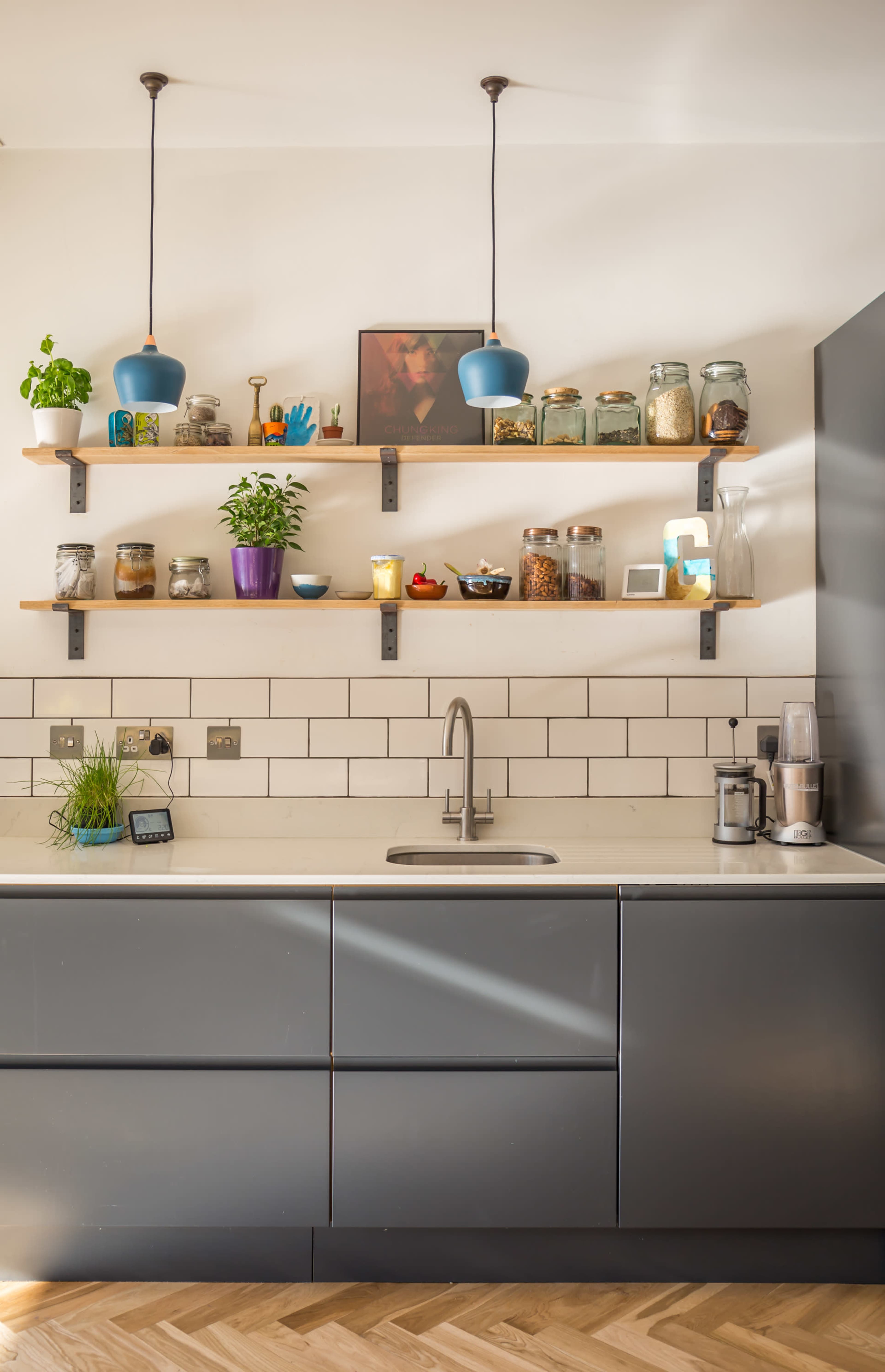 A modern kitchen features gray cabinetry, open wooden shelves filled with jars and plants, and a stainless steel sink.