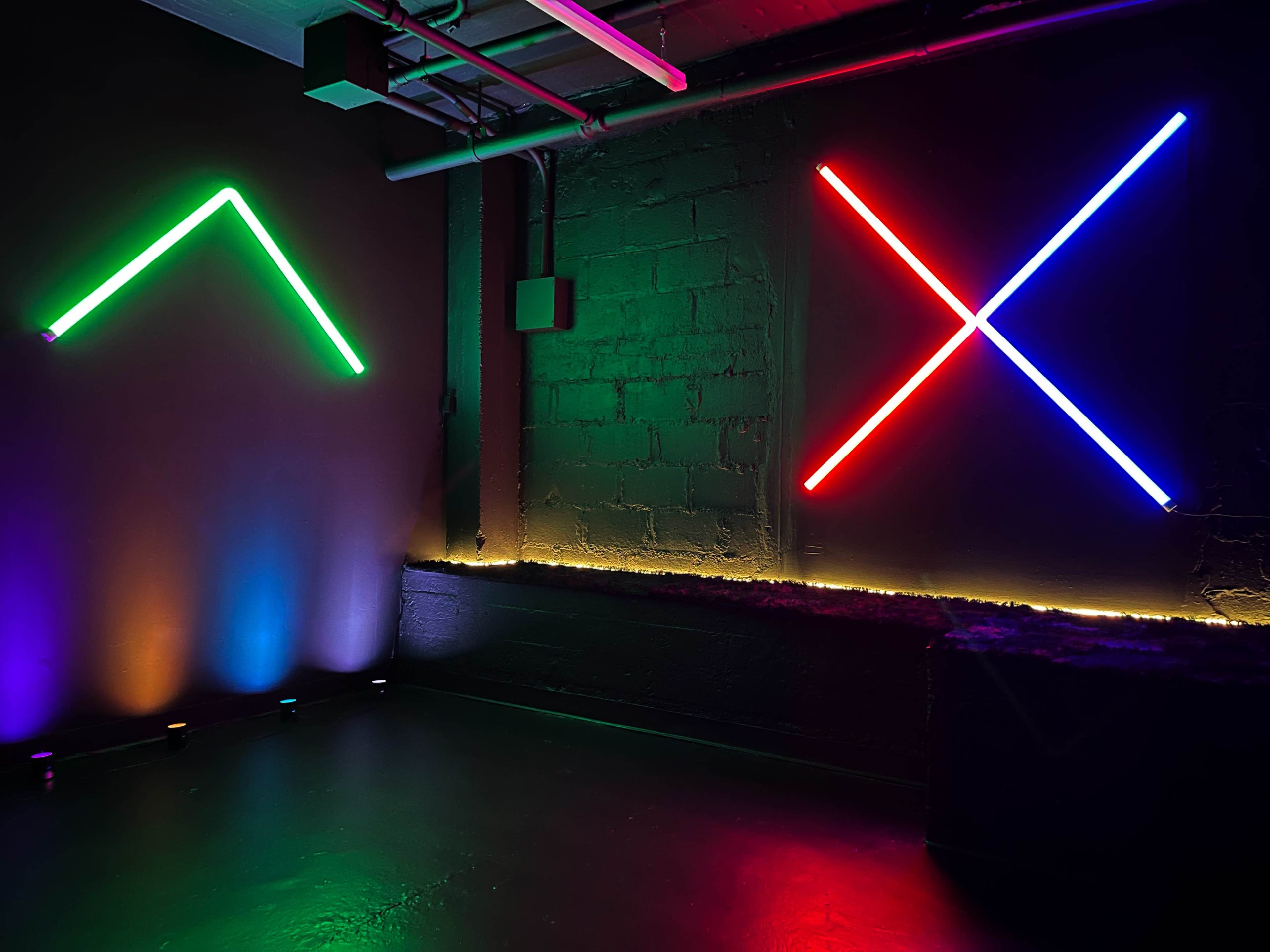 A dark room with illuminated neon light designs forming an upward triangle and an "X" shape on opposite walls.