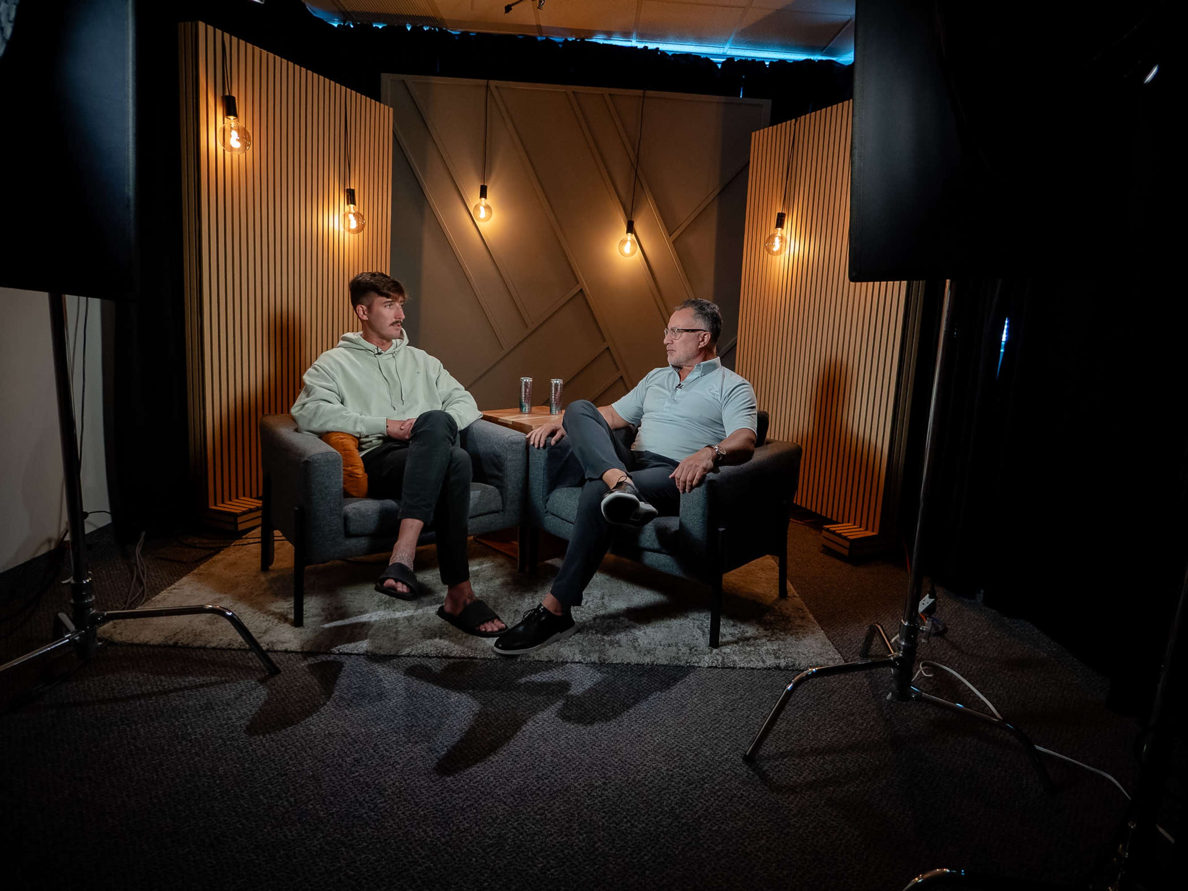 Two men sit in gray armchairs on a rug in a well-lit studio, discussing while holding drinks, with wooden paneling and light fixtures in the background.