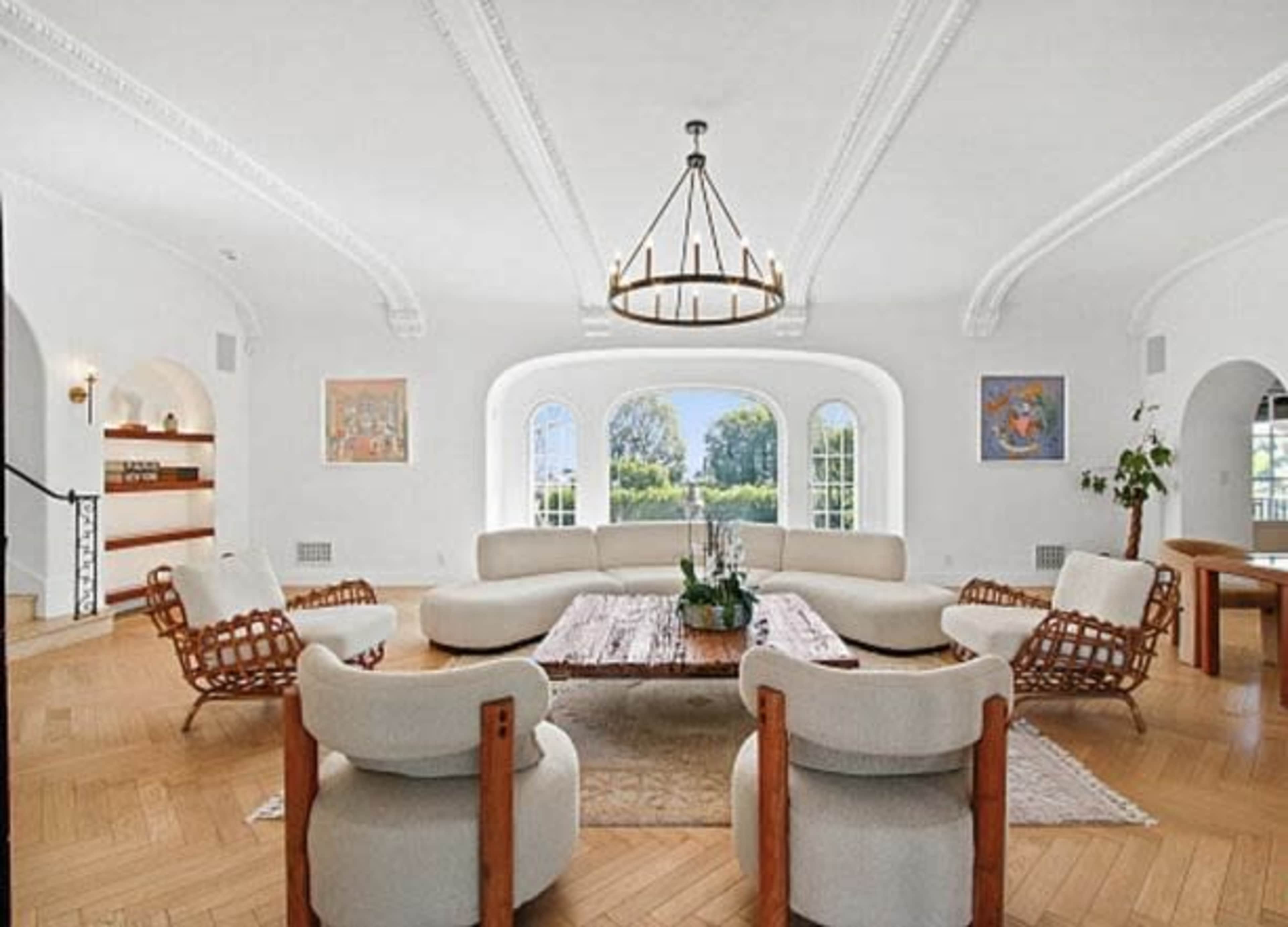 The image shows a spacious living room with a round wooden coffee table, white sofas, and large arched windows overlooking greenery.