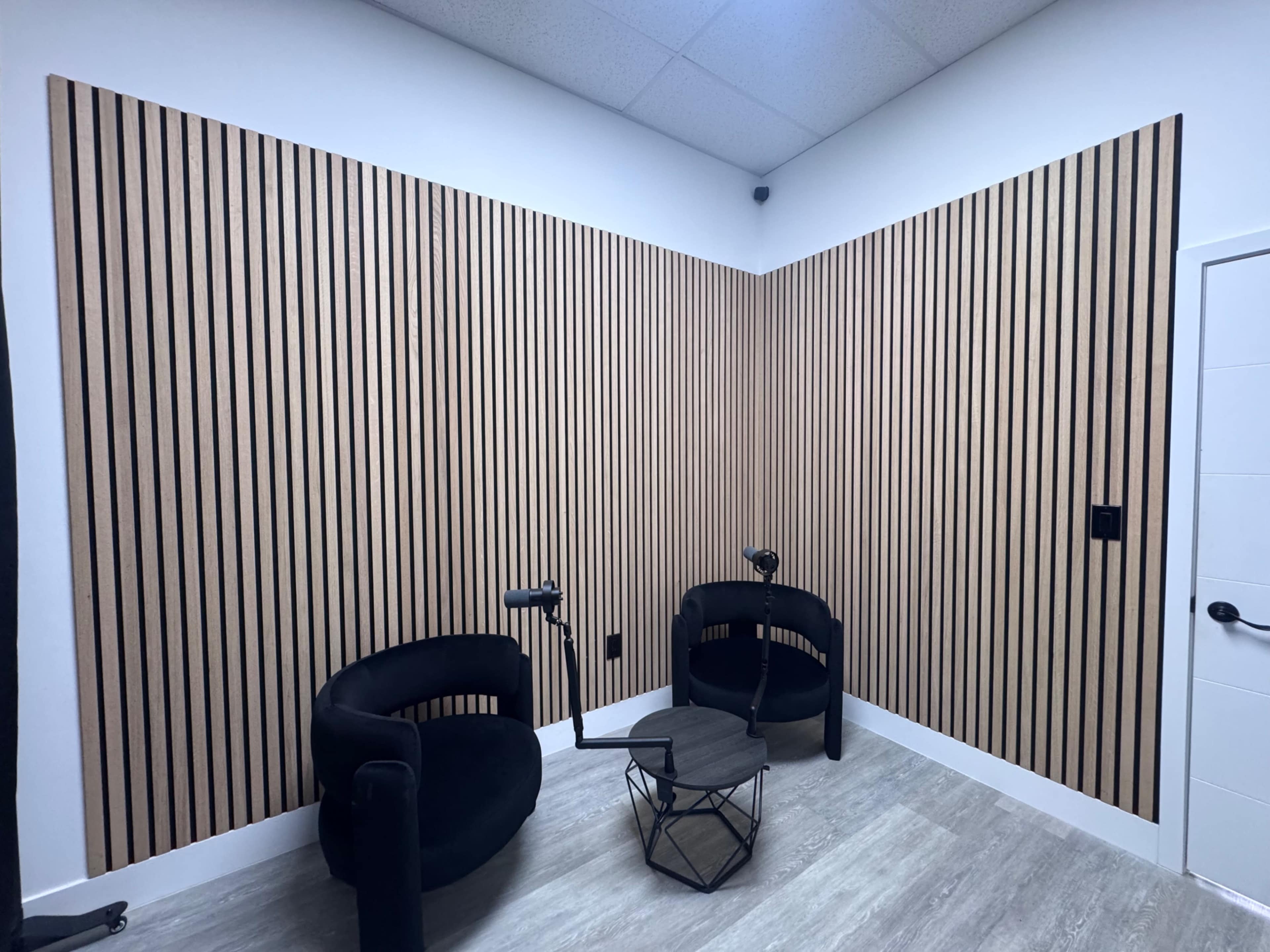 The image shows a corner of a room with vertical wooden slat walls, featuring two black chairs and a small round table.