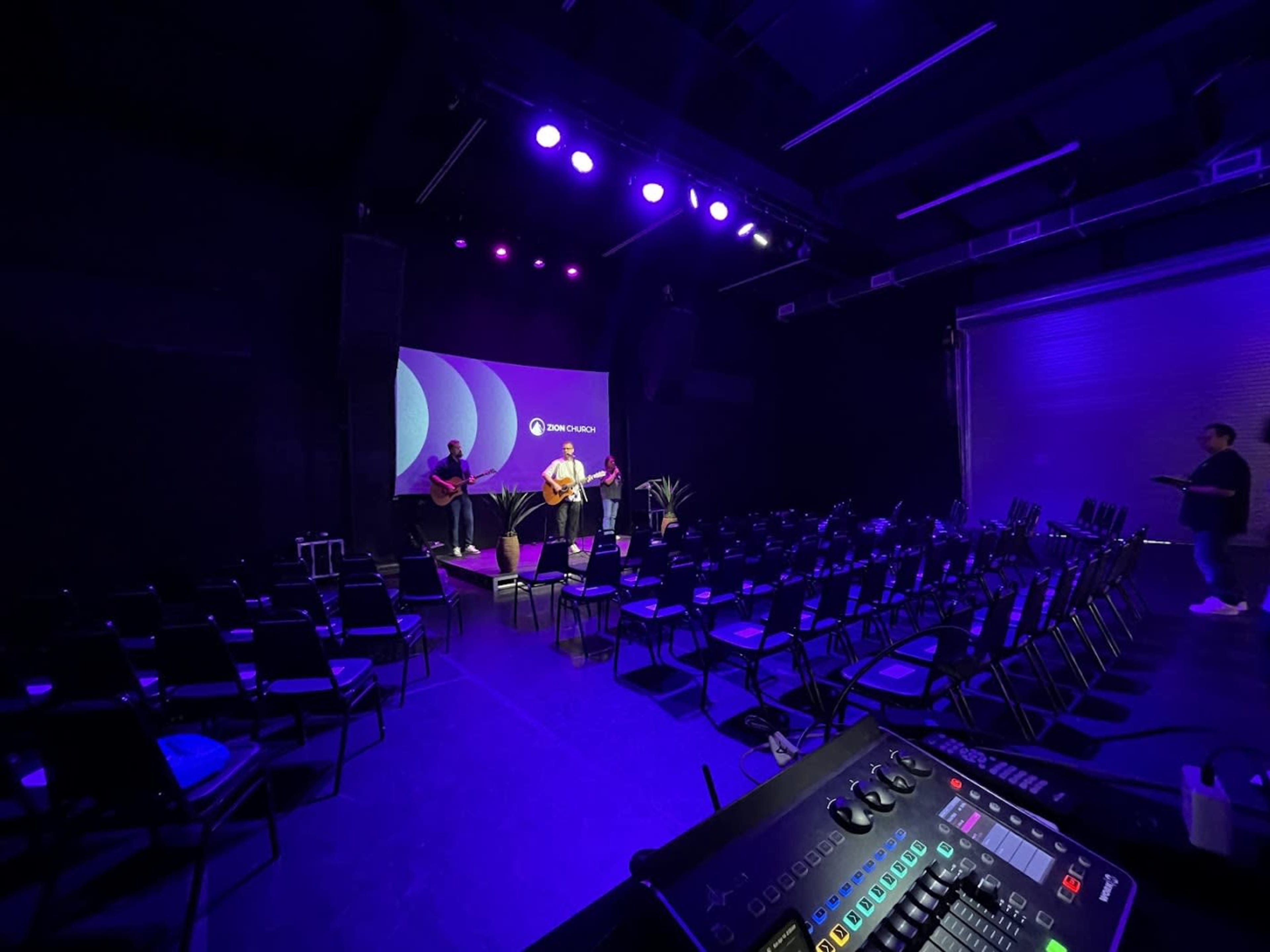 A musician plays guitar in a dimly lit performance space with chairs arranged for an audience and a control panel in the foreground.