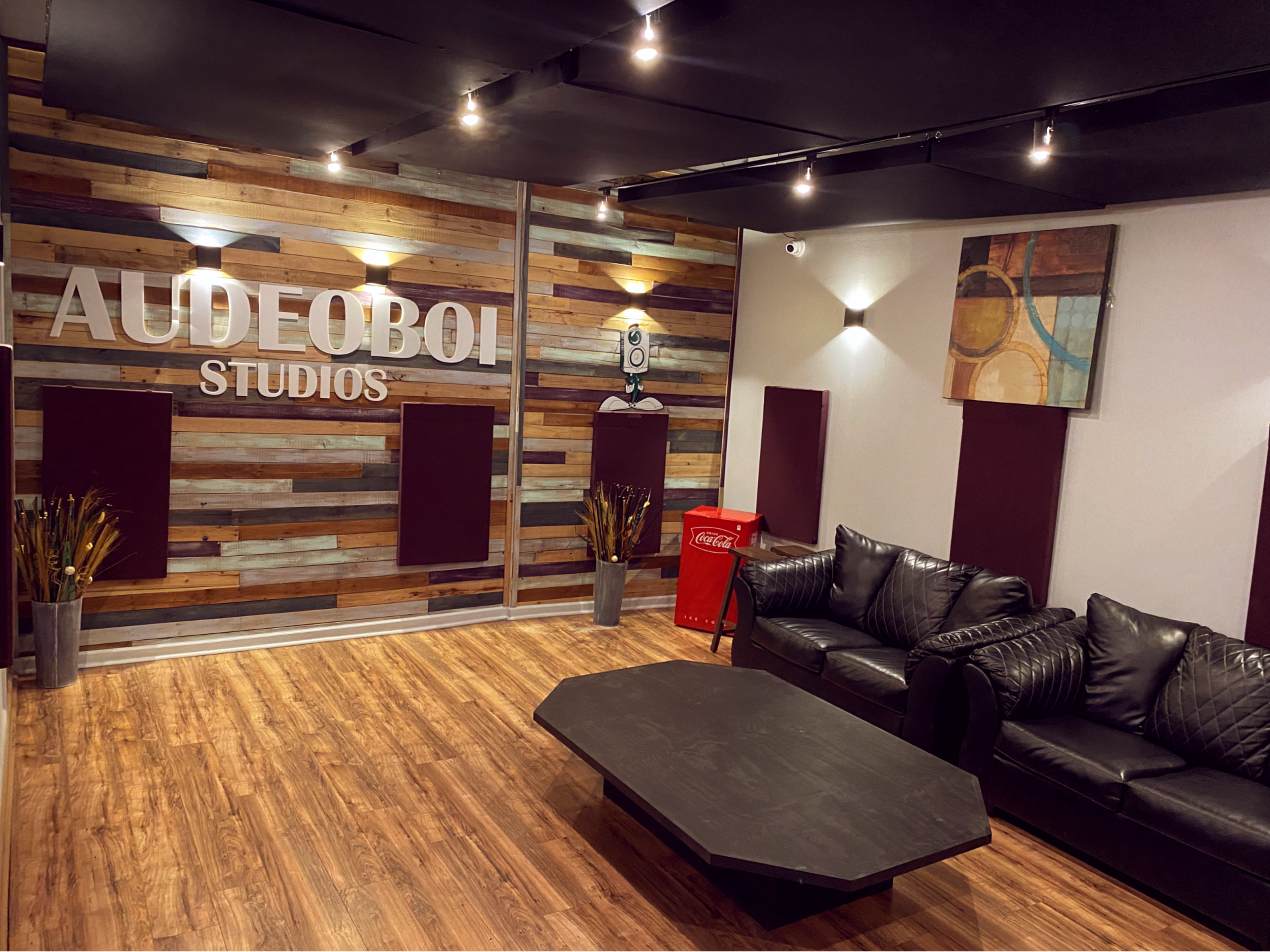 A modern studio lounge with wooden paneling on the walls, a black coffee table, a black leather sofa, and a Coca-Cola vending machine in the corner.