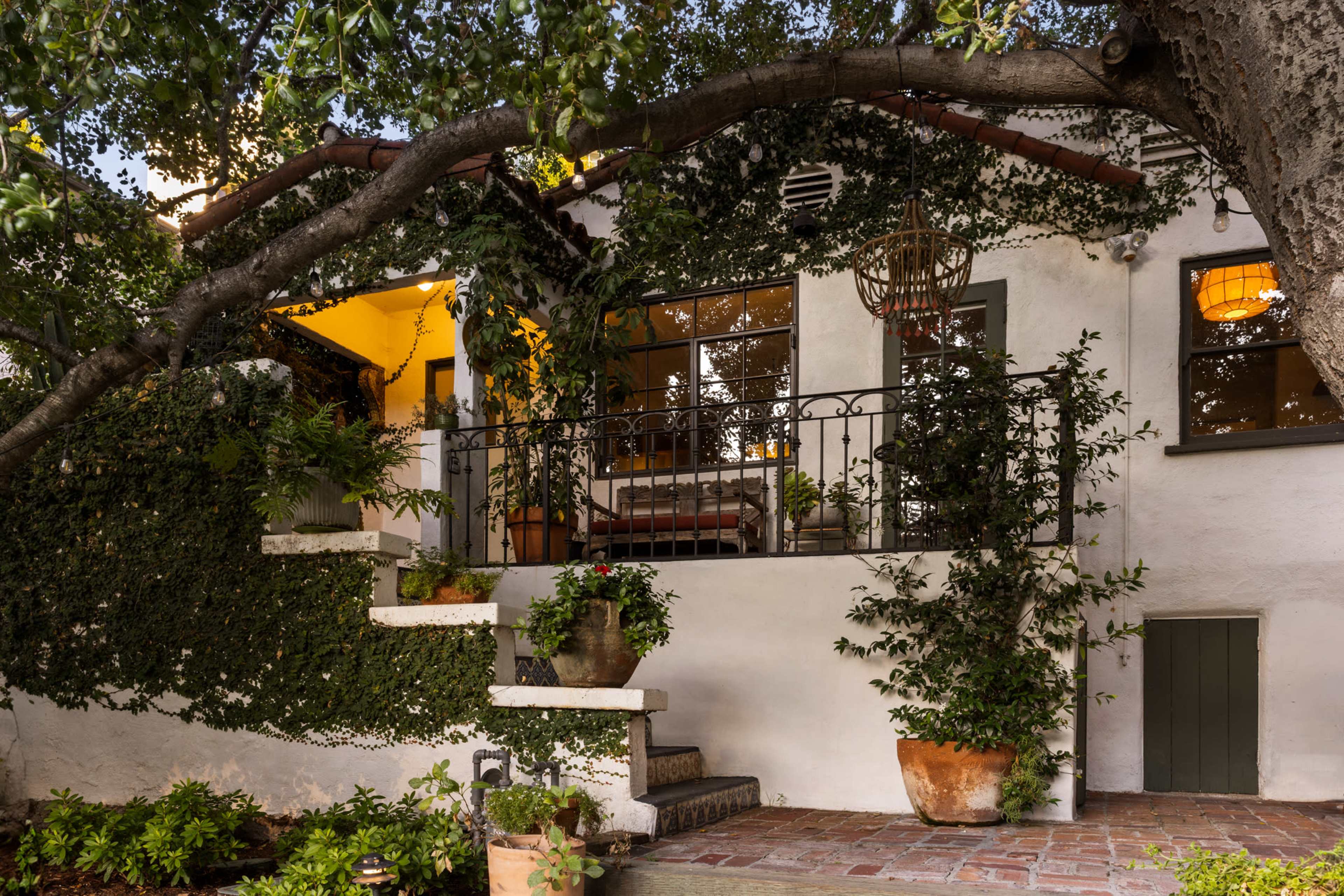 The image shows a quaint house with a covered porch, surrounded by lush greenery and potted plants, featuring a staircase leading to the entrance.