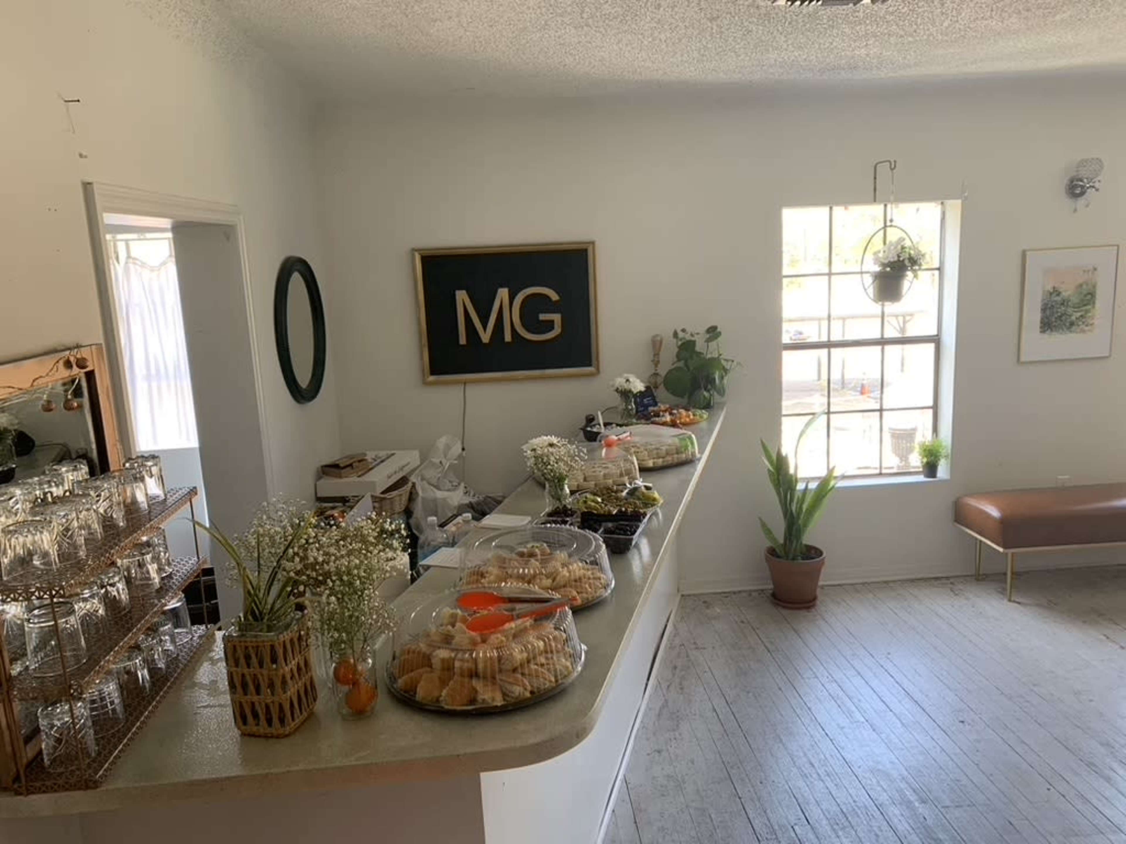 A brightly lit café interior features a counter displaying an array of pastries and a wall adorned with a large sign reading "MG."