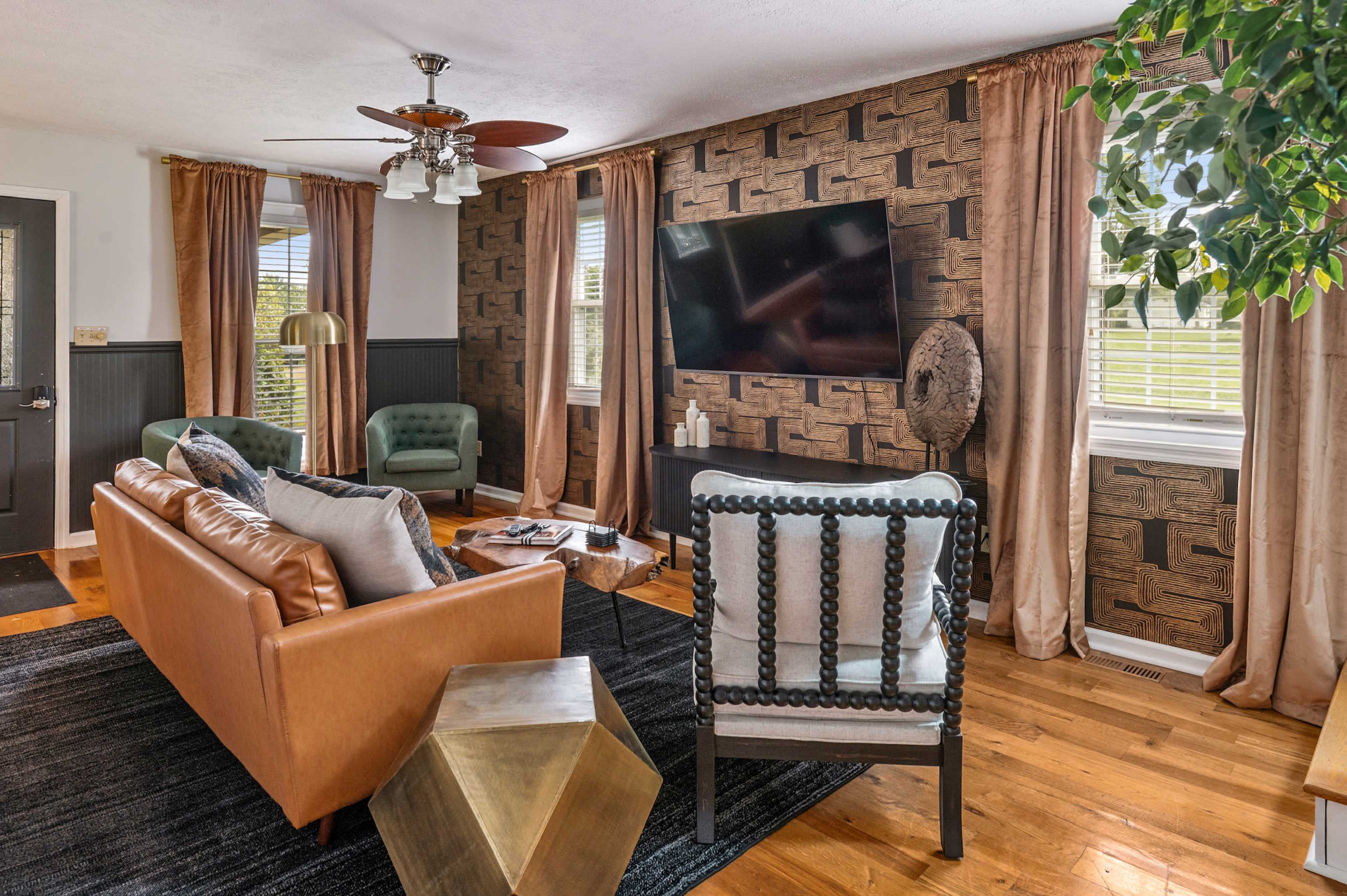 The image shows a cozy living room featuring a brown leather sofa, black and white accent chairs, a coffee table, and a flat-screen TV mounted on a patterned wall, complemented by large windows and light-colored drapes.