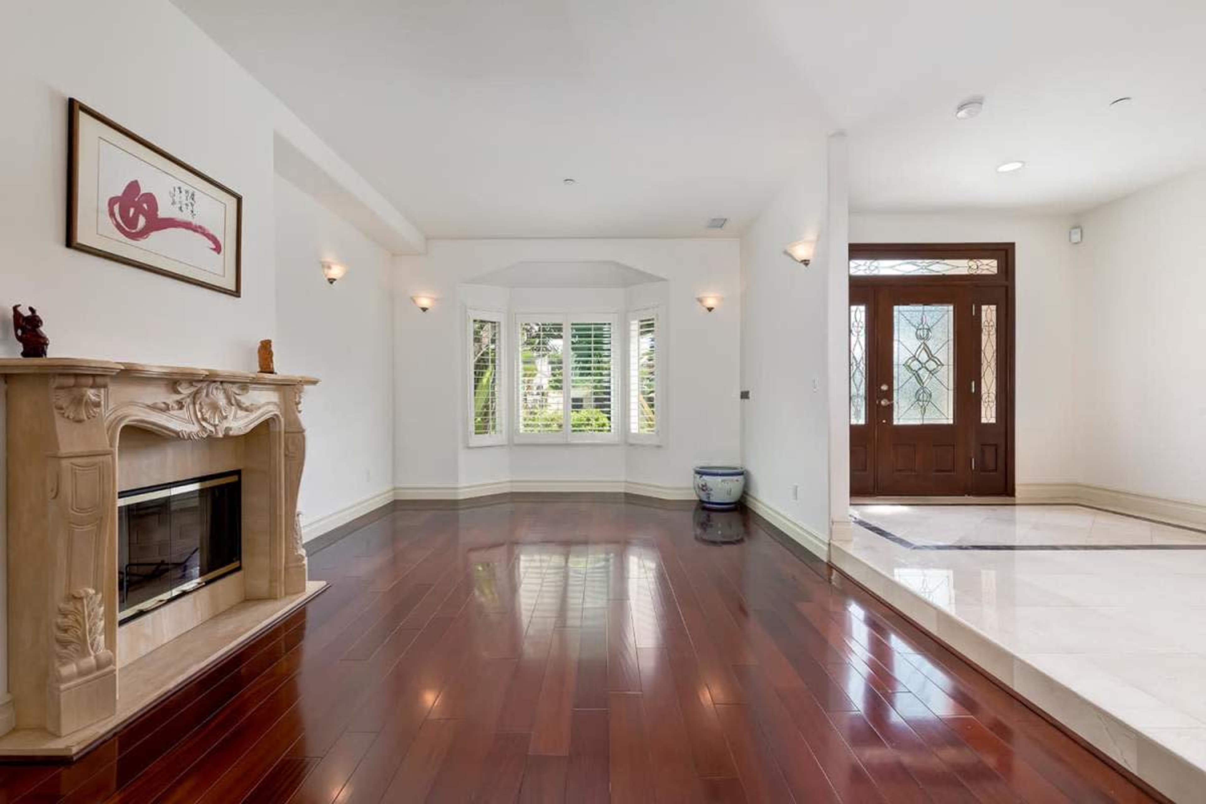 The image shows a spacious interior with hardwood flooring, a decorative fireplace, and large windows, leading to a front door with intricate glass design.