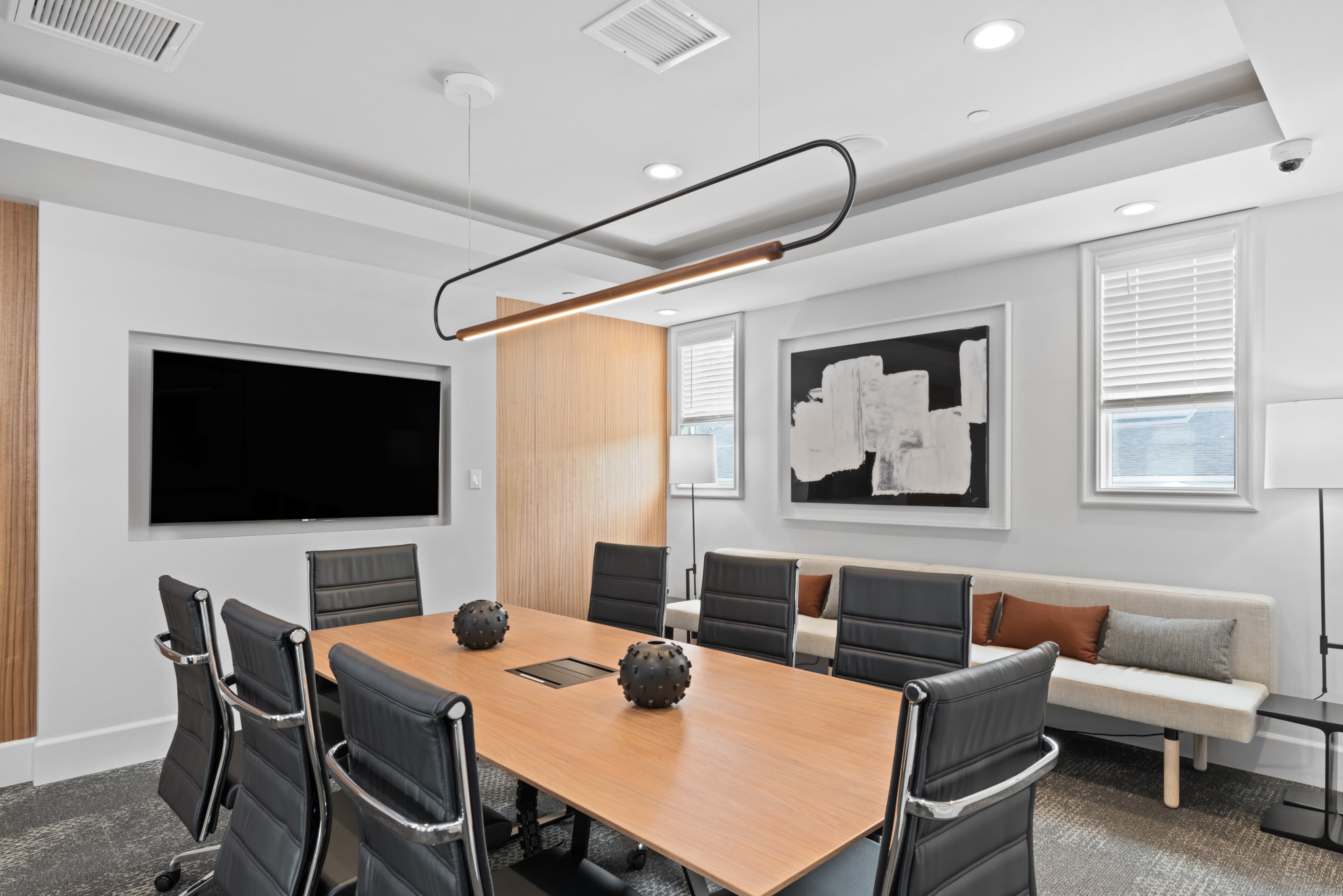 A modern conference room featuring a long wooden table surrounded by black leather chairs, a large screen on the wall, and a minimalist artwork.