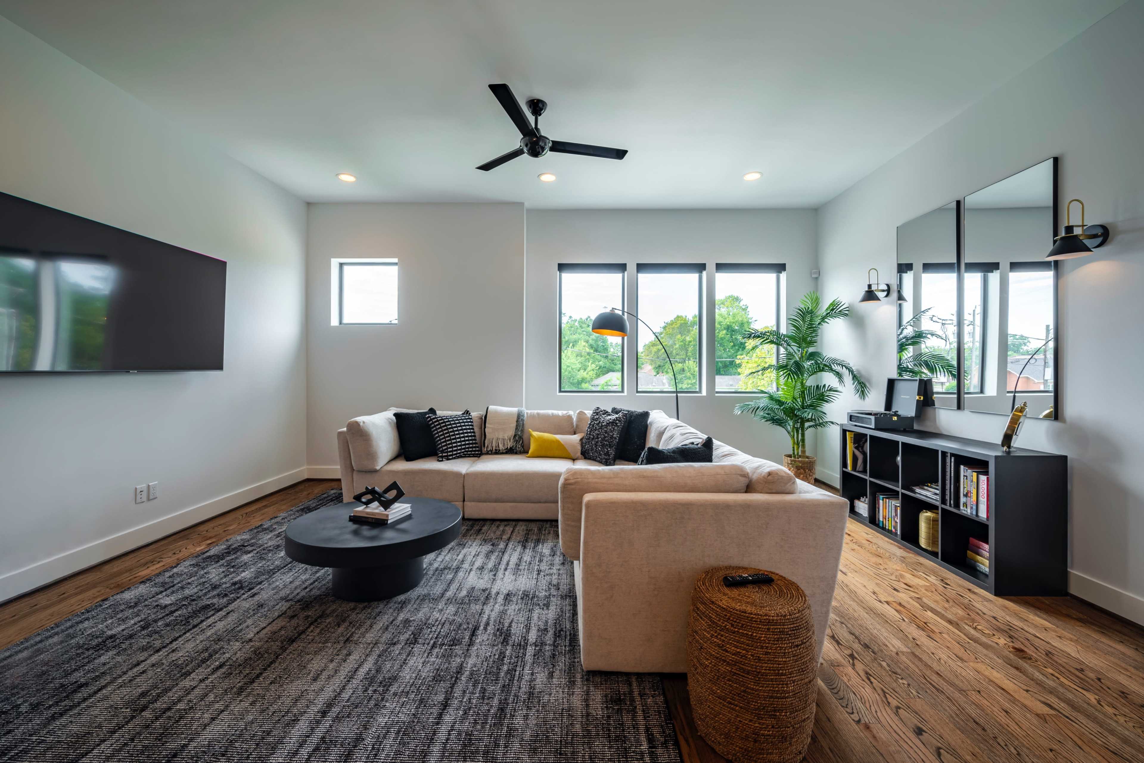 The living room features a large sectional sofa, a round coffee table, and a wall-mounted television, with windows providing natural light and a nearby bookshelf.