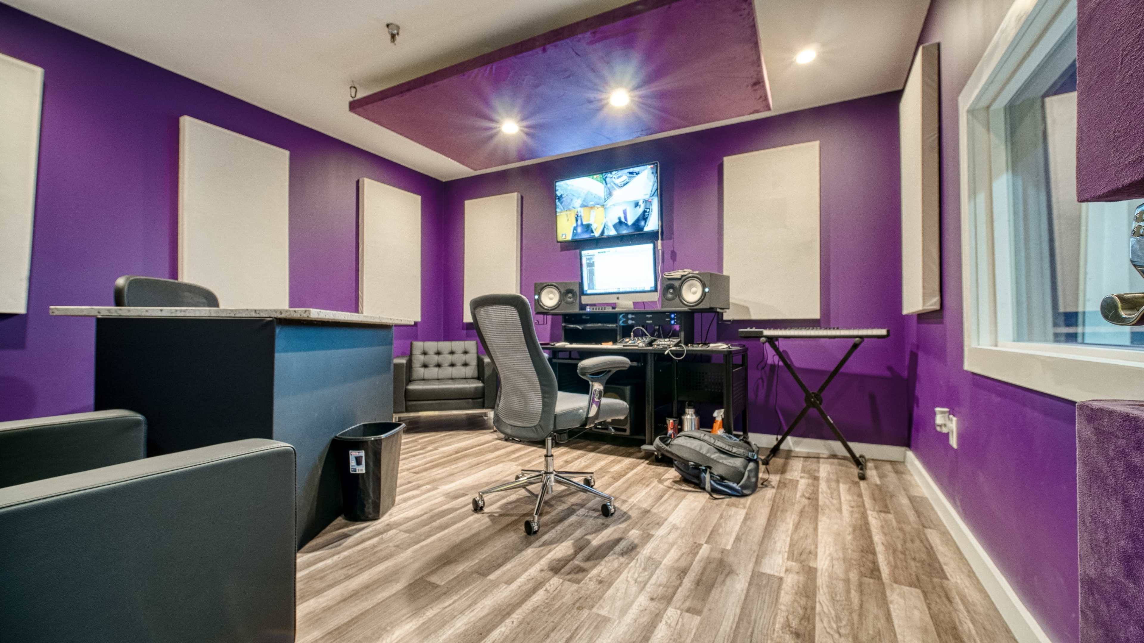 A music production studio with purple walls, a central desk with monitors, a keyboard, and a seating area.