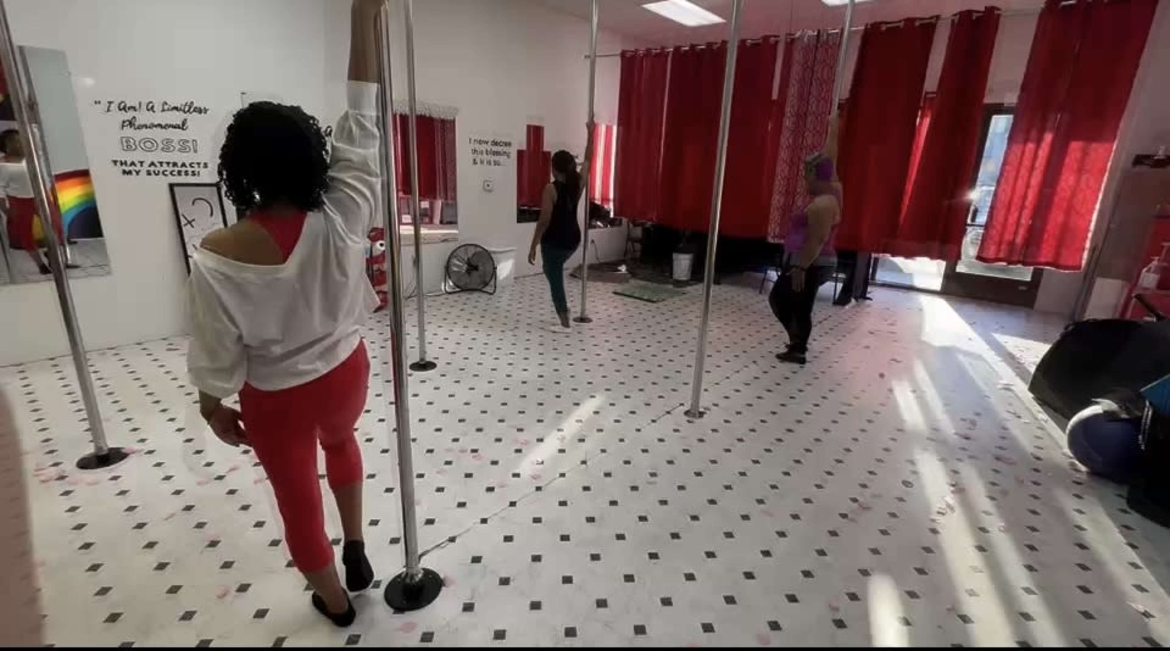 A group of people practices pole dancing in a studio with mirrors and red curtains.