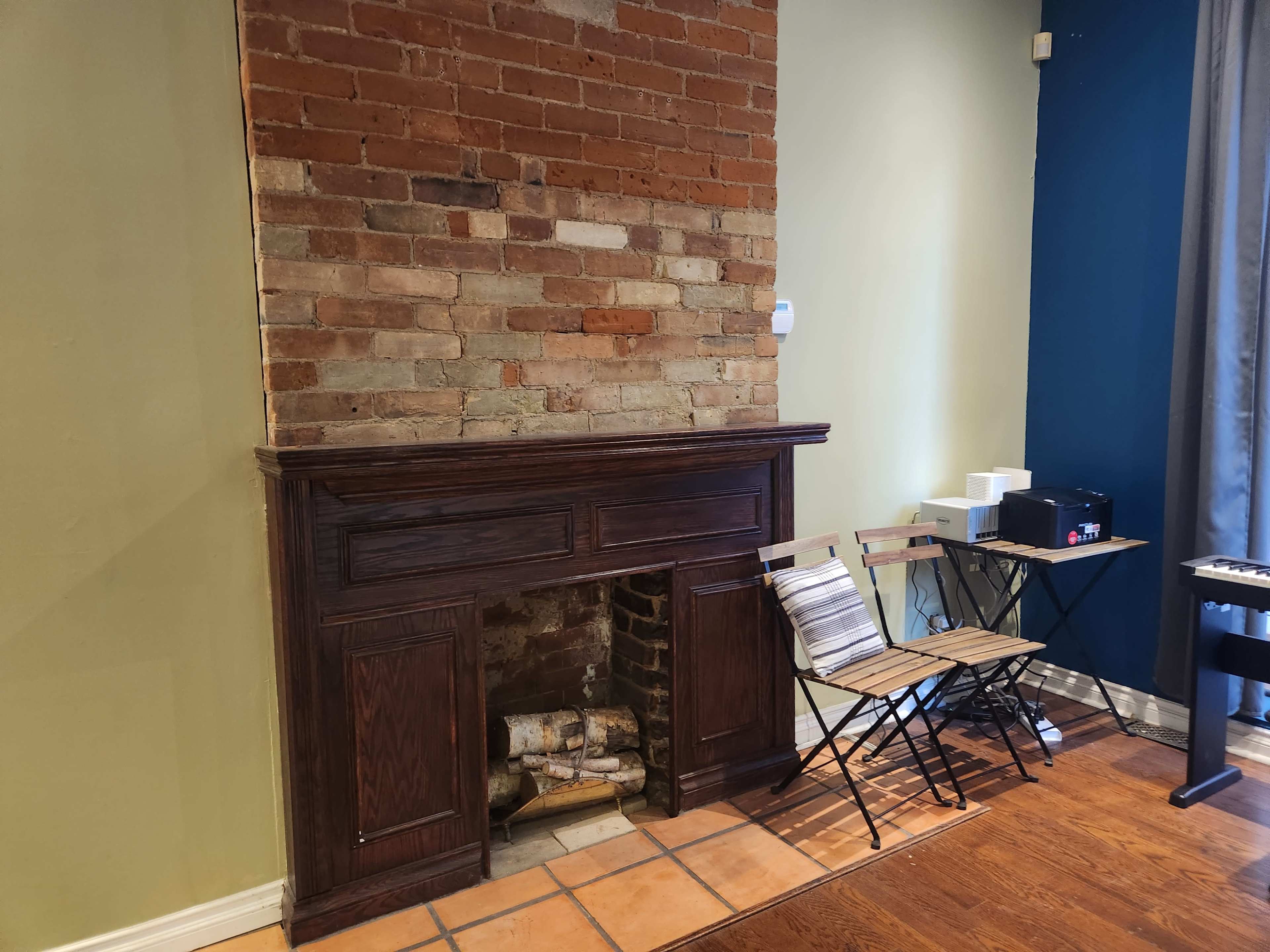 A brick wall with a wooden mantel and an empty fireplace, accompanied by a small wooden table and chair set beside it.
