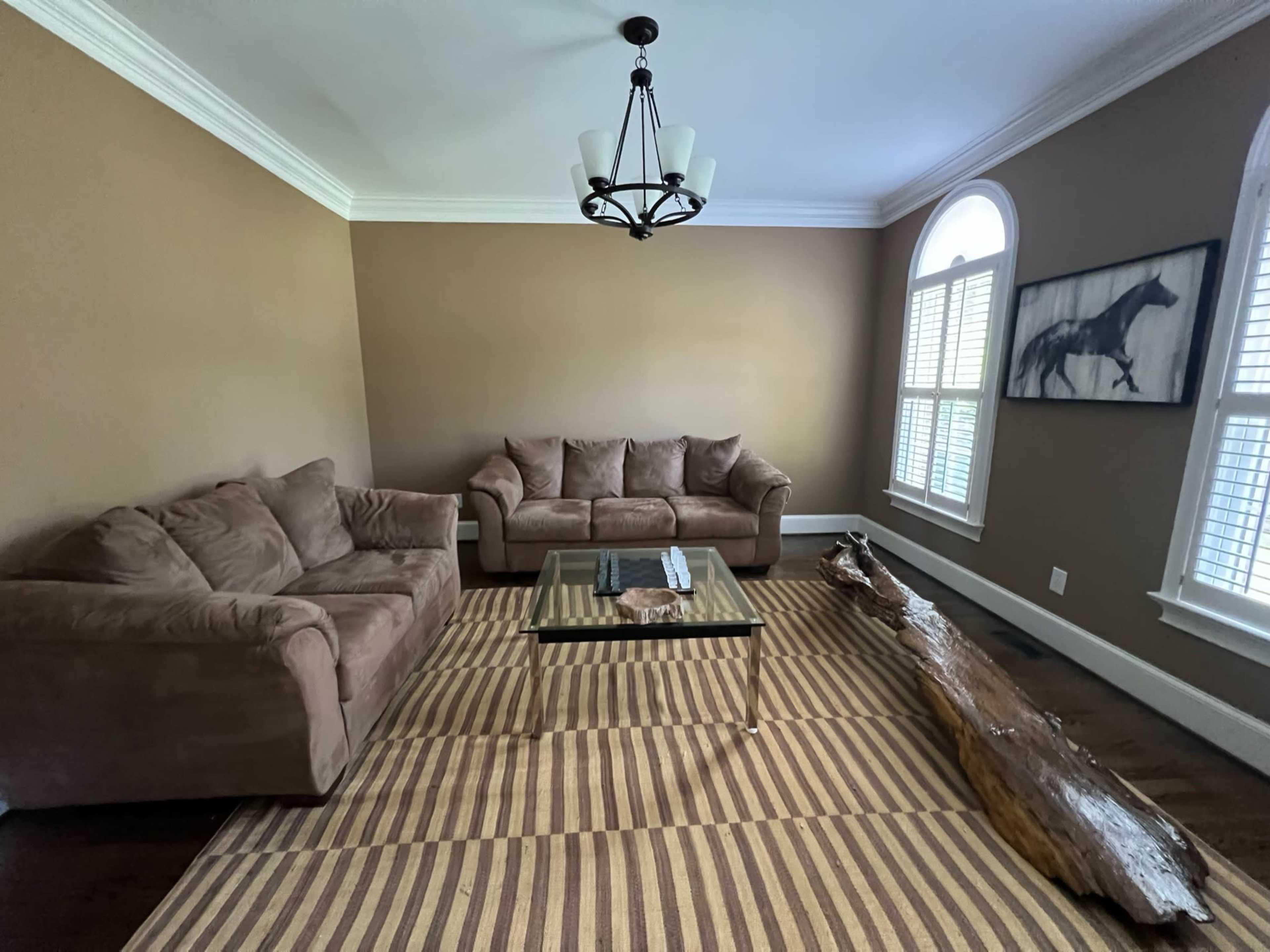 A living room with two brown sofas, a glass coffee table, a decorative wooden log, and large windows with white shutters.