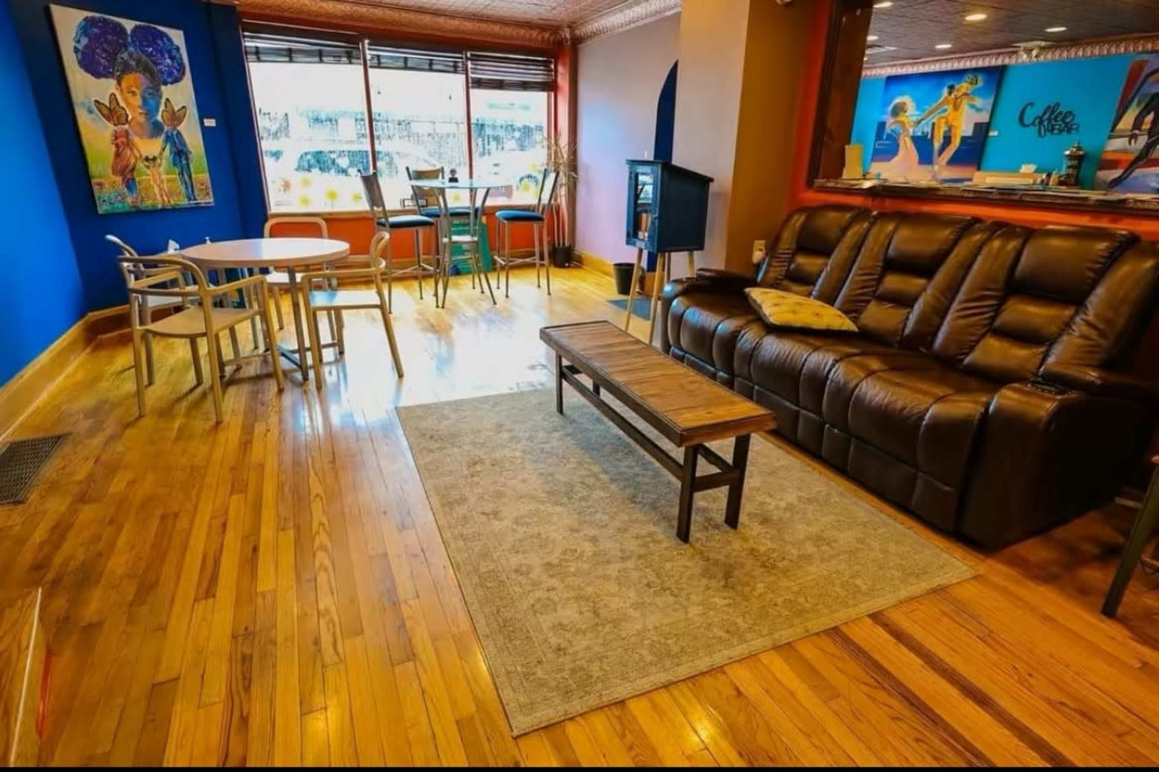 The image shows a cozy café interior with wooden flooring, a brown leather couch, a wooden coffee table, and a mix of seating arrangements including small tables and chairs.