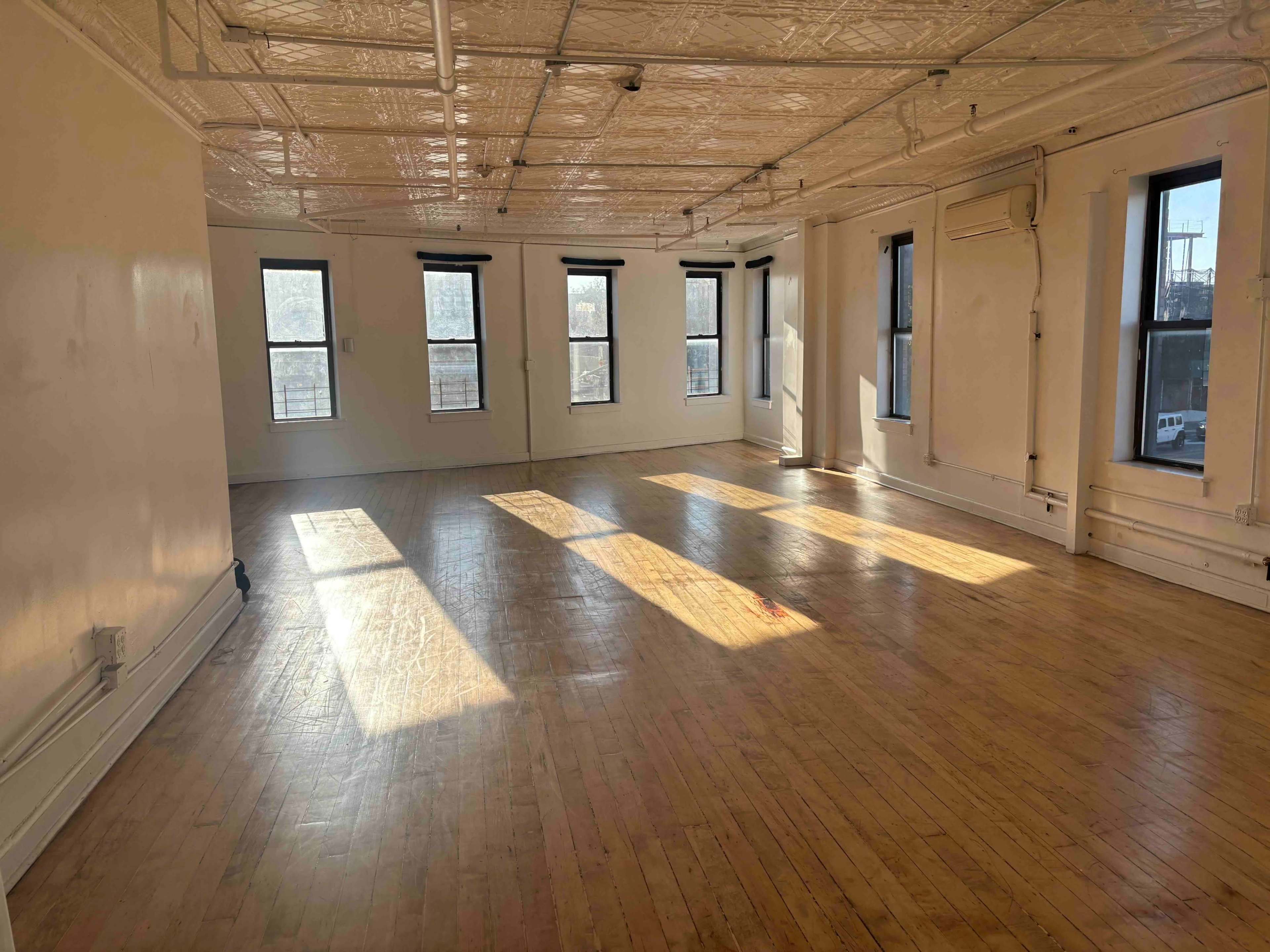 The image shows a spacious, empty room with wooden floors and several windows allowing natural light to enter.