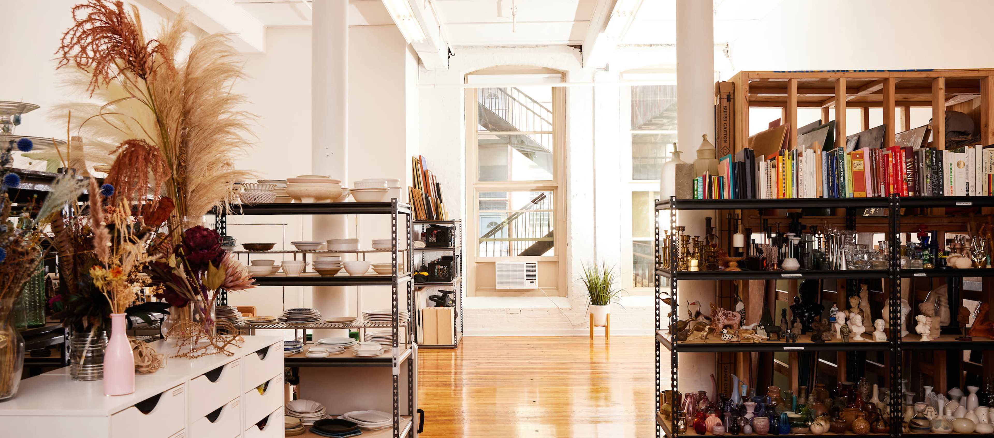 The image depicts a bright, open space filled with shelves of various home goods, including plates, books, and decorative items, with large windows allowing natural light to enter.