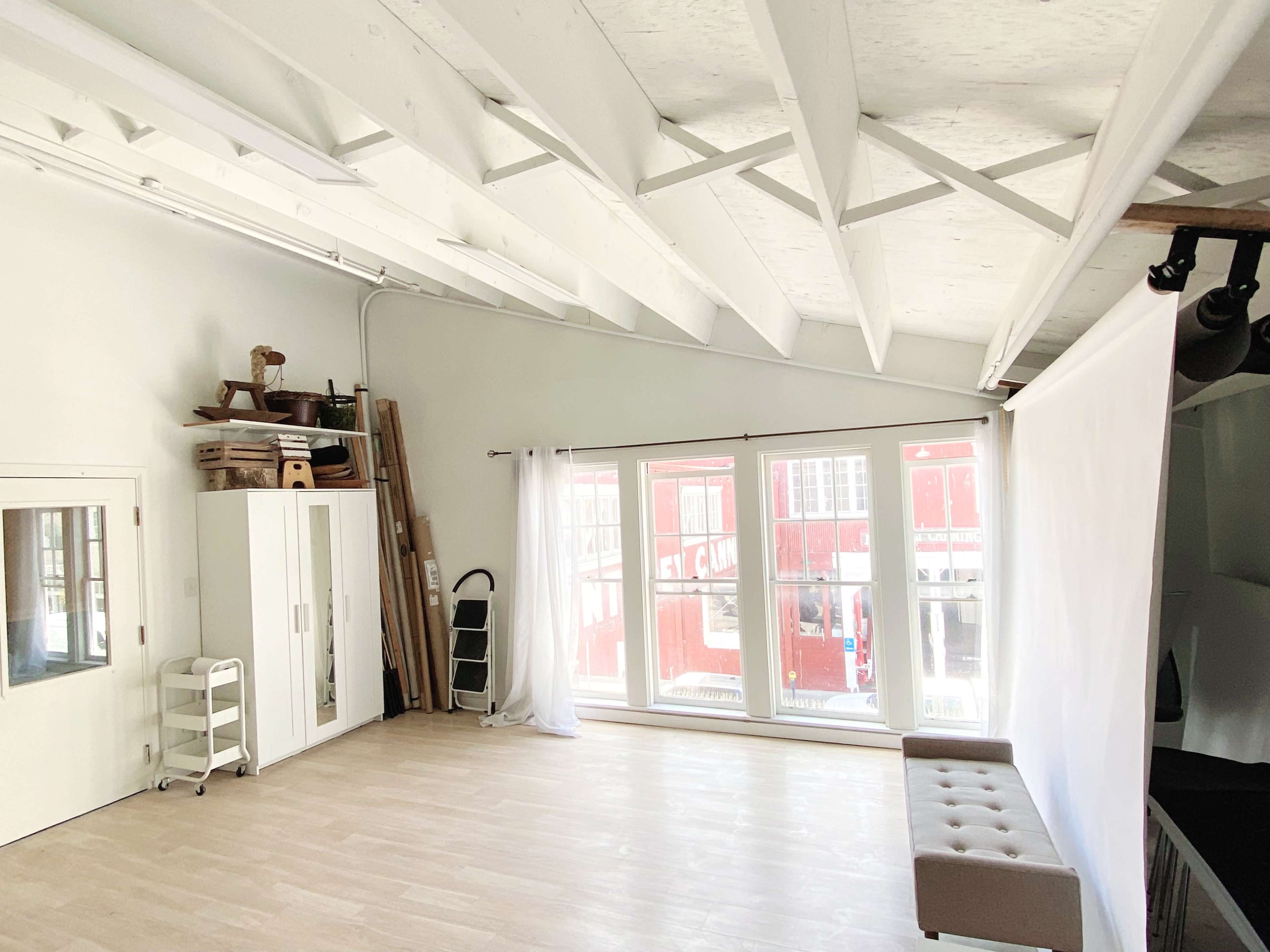 A spacious, well-lit room with high, white-beamed ceilings, featuring a large window, a curtain backdrop, and minimal furniture.