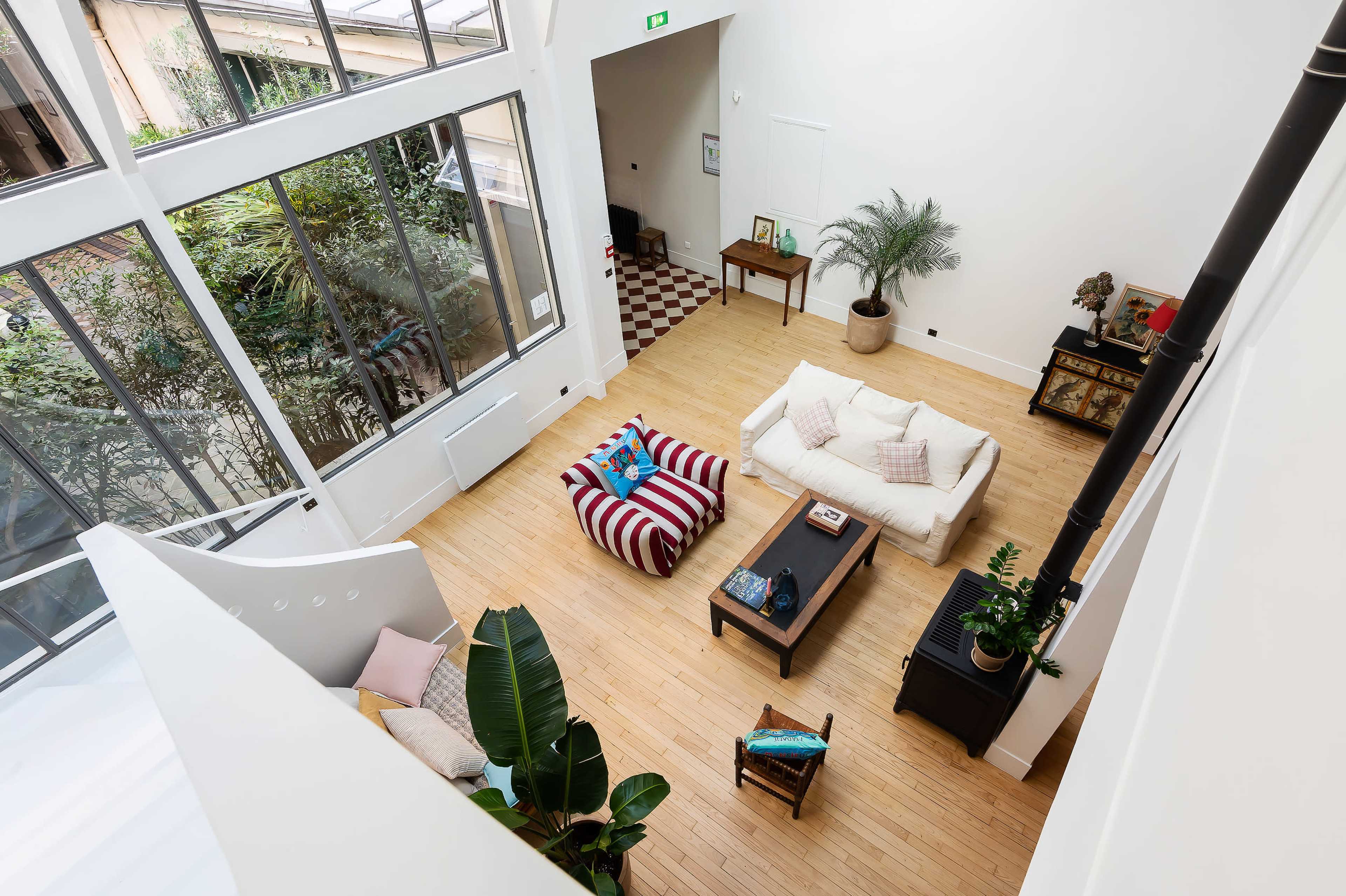 The image shows an airy, modern living room with large windows, wooden flooring, and a mix of furniture including a striped sofa, a coffee table, and decorative plants.