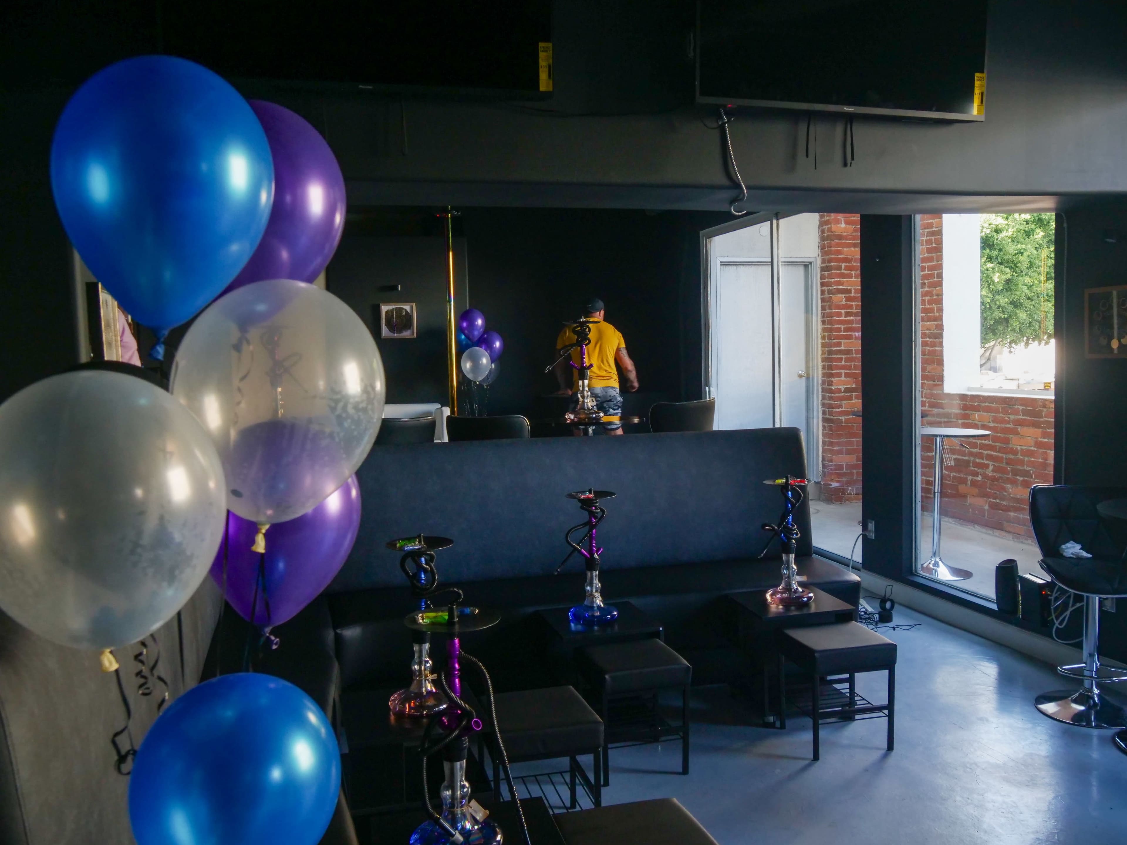 A lounge area with black seating, colorful balloons, and hookahs, while a person adjusts something in the background.