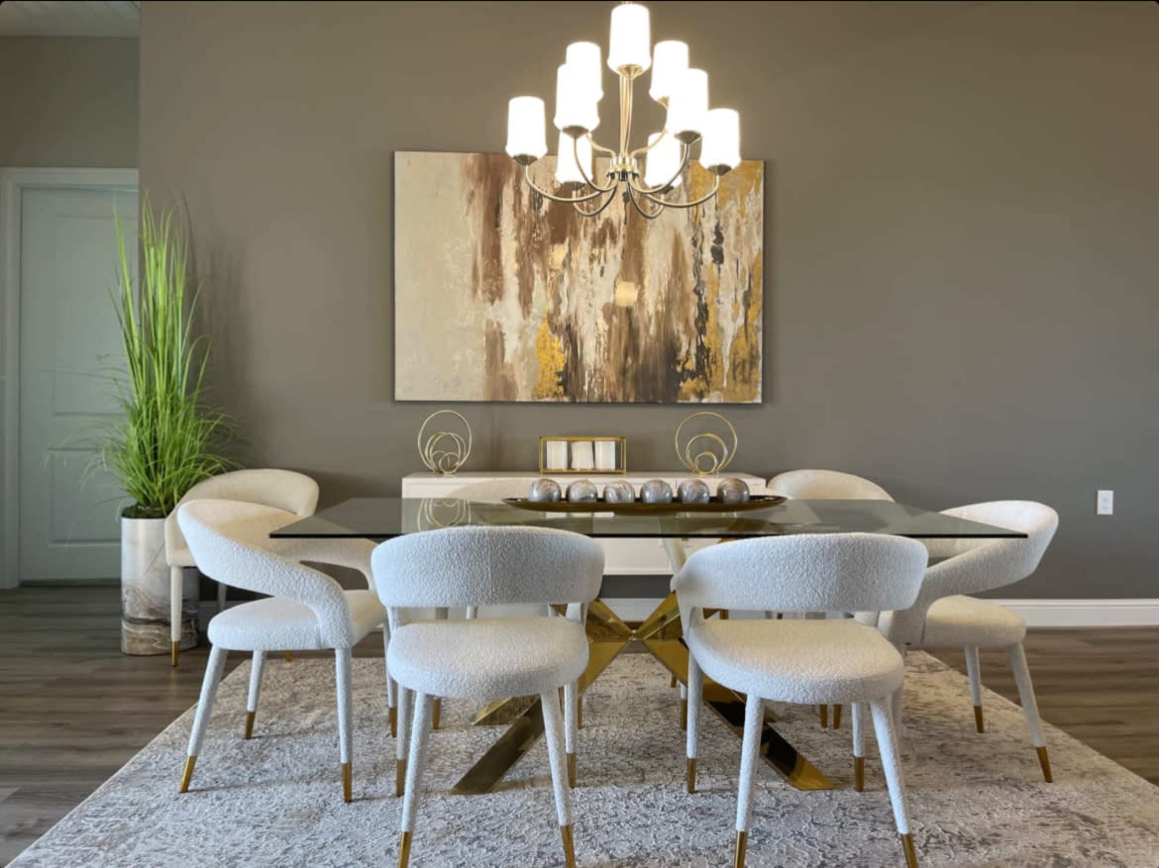 A modern dining room features a glass table surrounded by white upholstered chairs, a large painting on the wall, and a chandelier above.