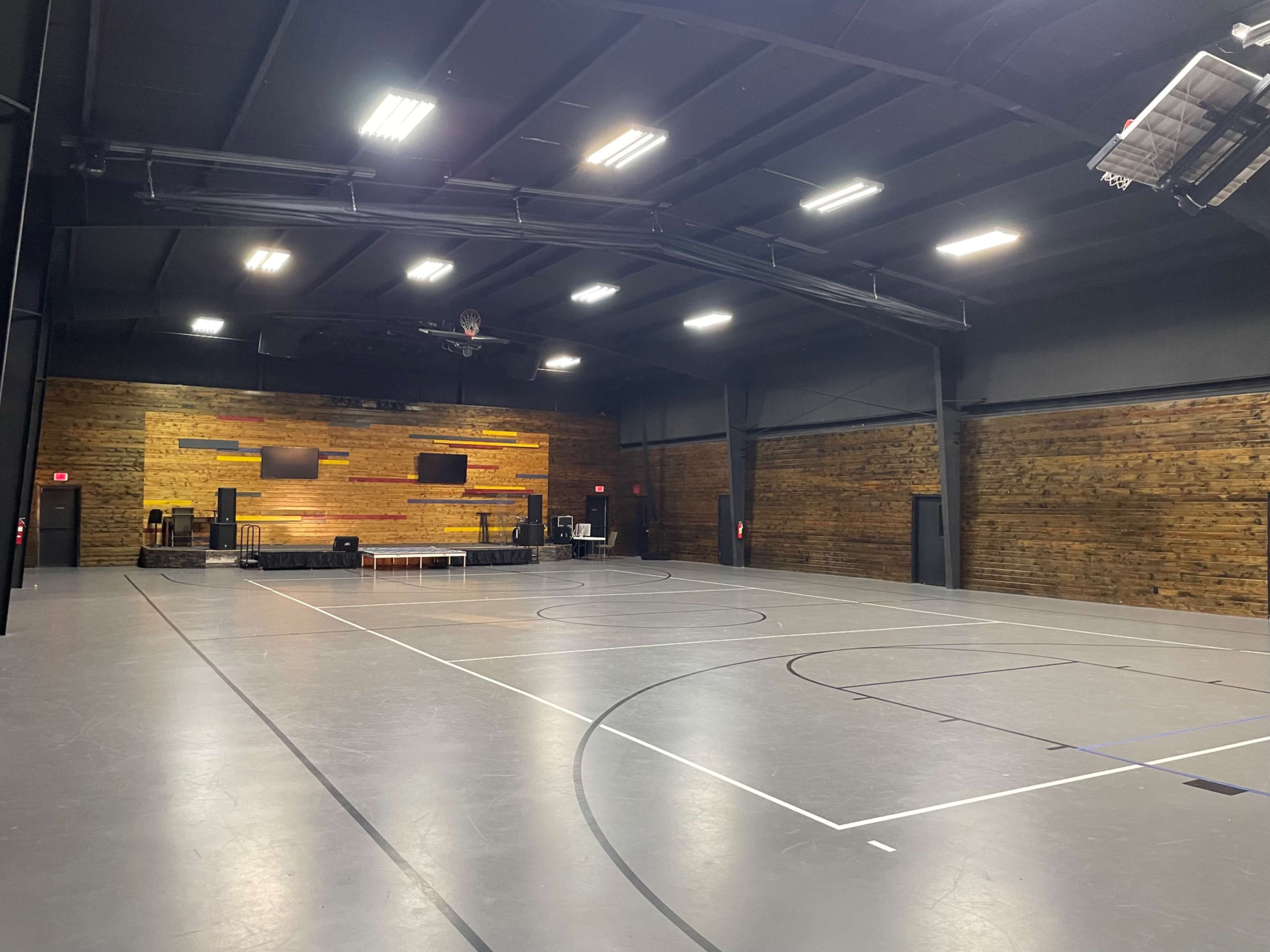 The image shows an indoor sports facility with a wooden accent wall, basketball court markings on the gray floor, and equipment positioned at one end.