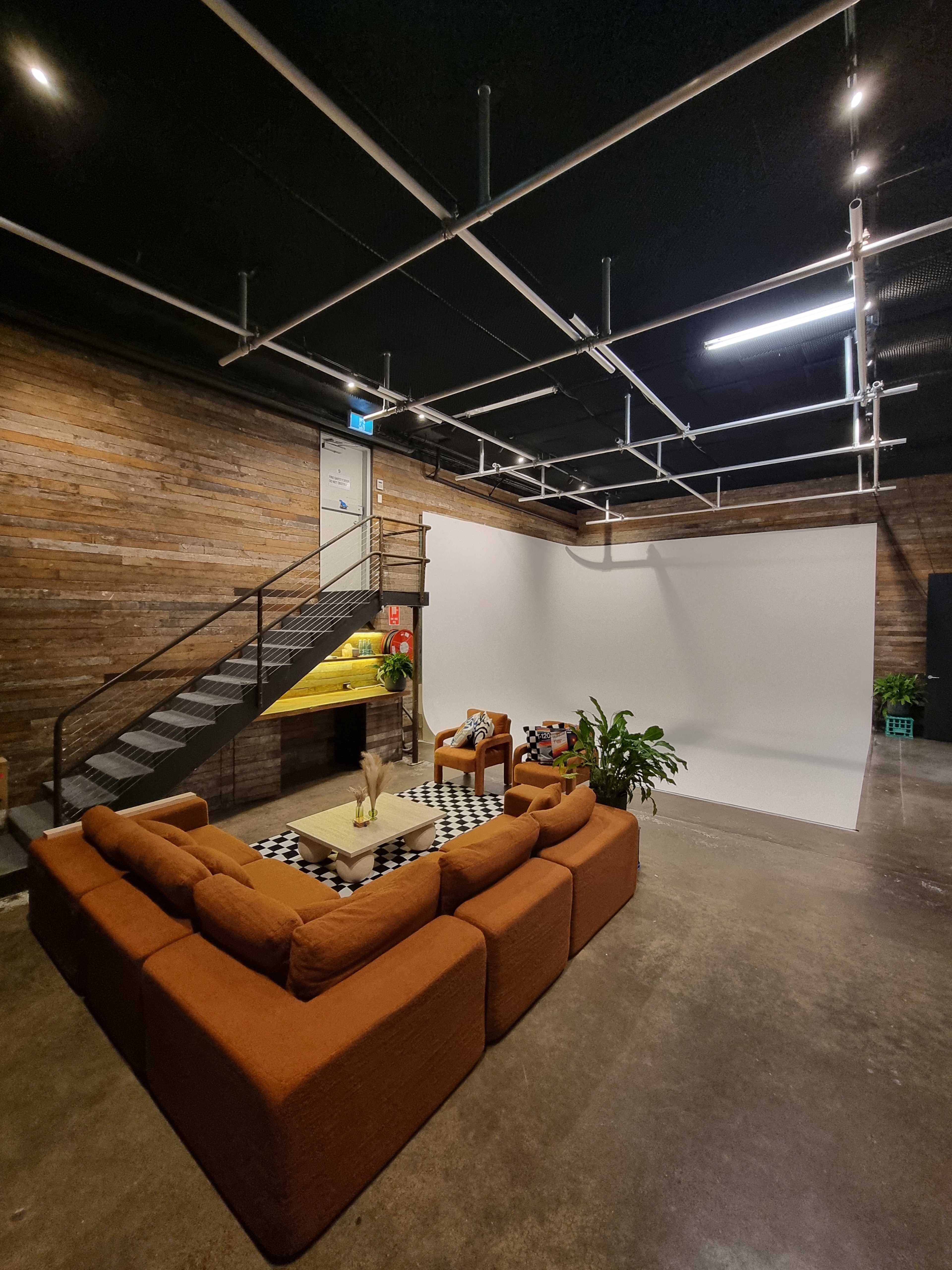 The image shows a spacious studio with a staircase, a large brown sectional sofa, and a white backdrop, along with plants and furniture arranged around a coffee table.